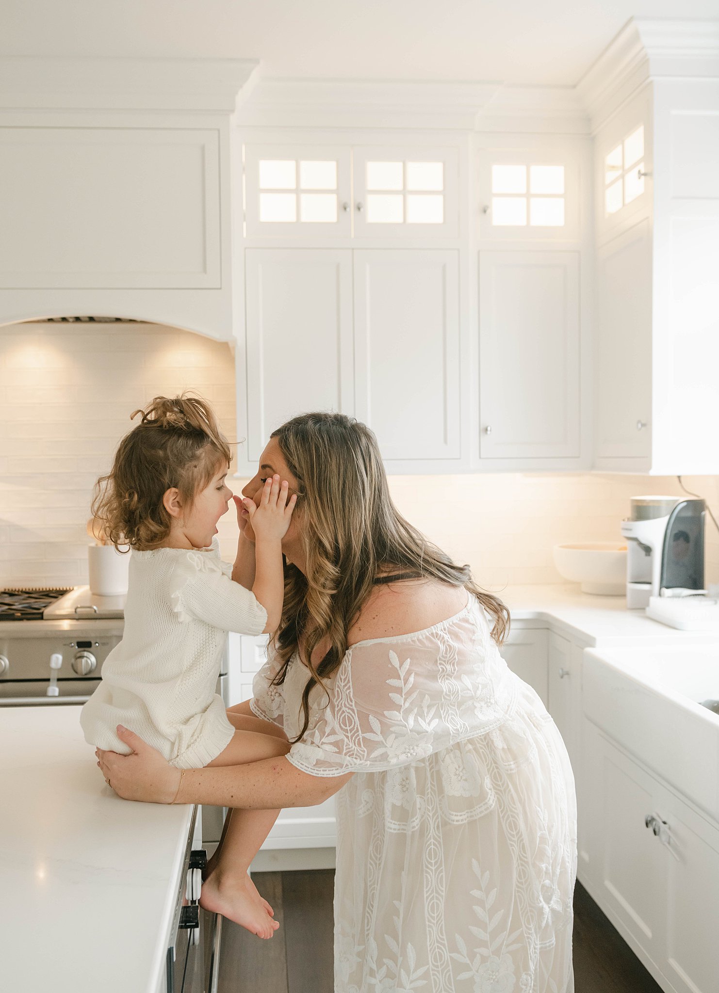 A young girl sits on the edge of a white kitchen island while playing with her mom in a white dress patting her face bellaboo lancaster