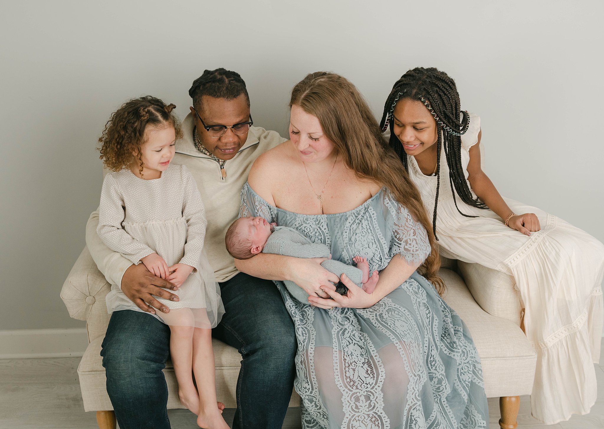 A family of five all sit together on a small tan couch in a studio looking down at the newborn baby sleeping in mother's arms
