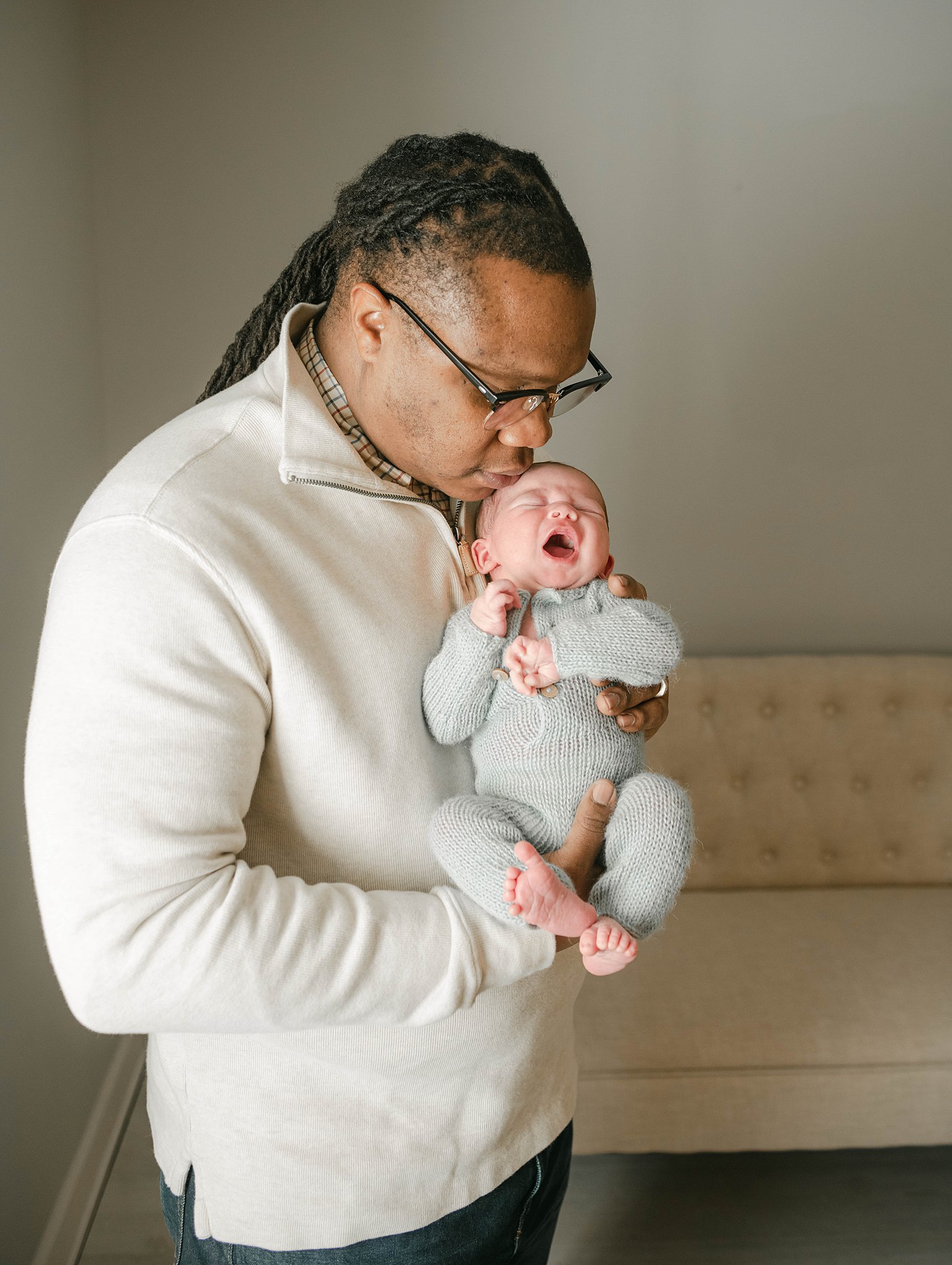 A father holds his yawning newborn baby to his chest wearing a tan sweater in a studio before we meet