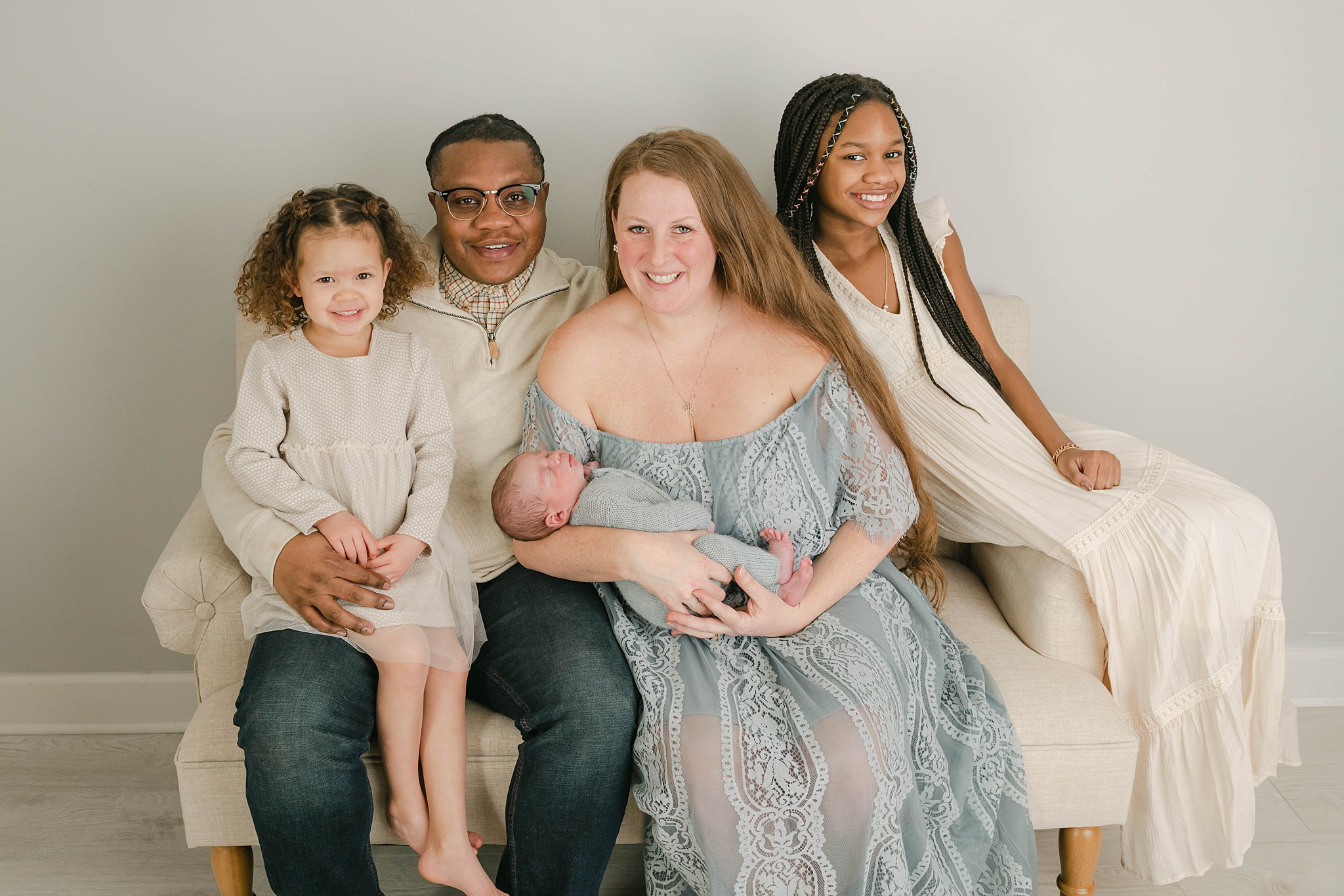 A family of five sits on a beige bench in a studio while mom holds the newborn baby