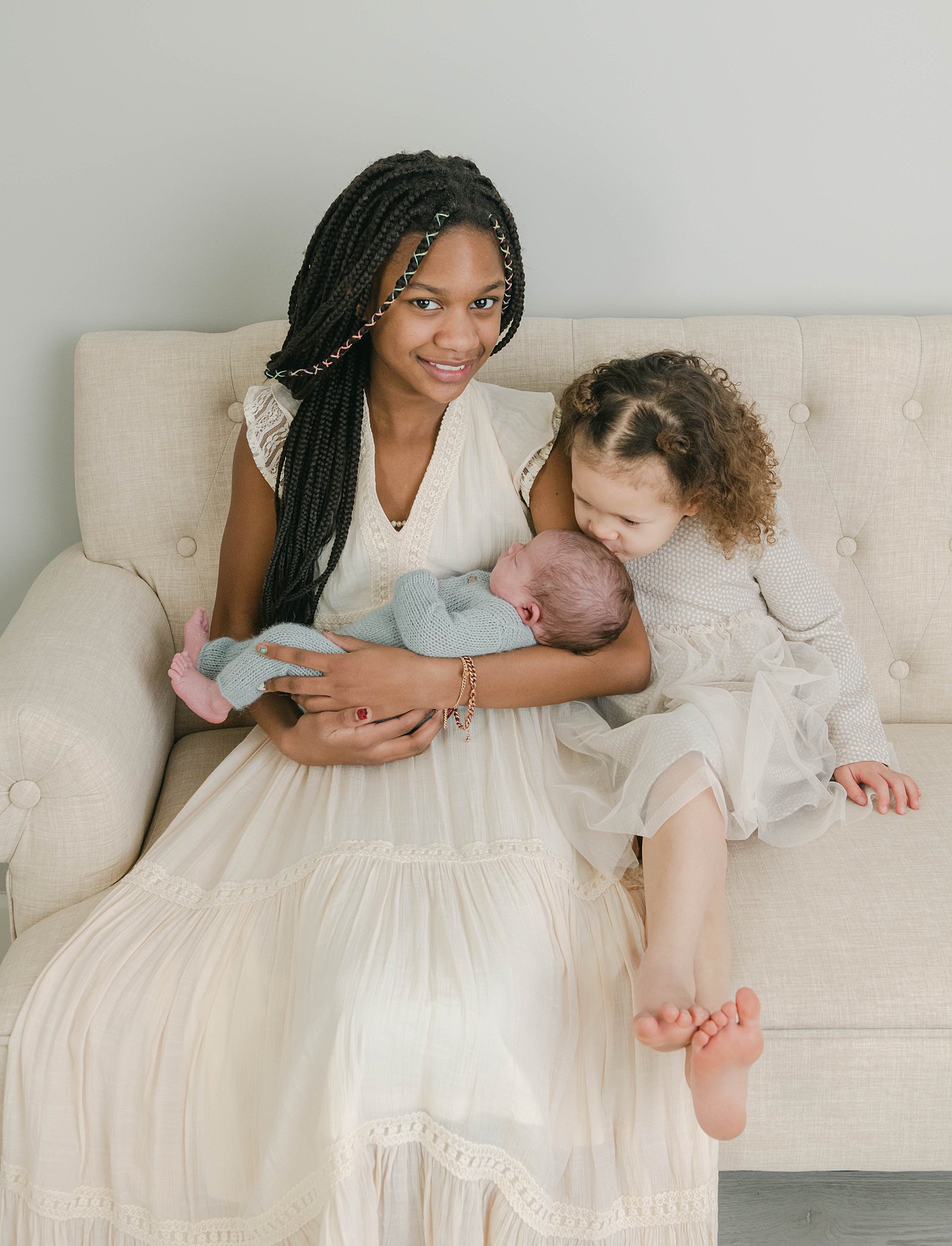 A teenager sits on a couch while holding her newborn baby brother while her toddler sister kisses the newborn's head the nesting house