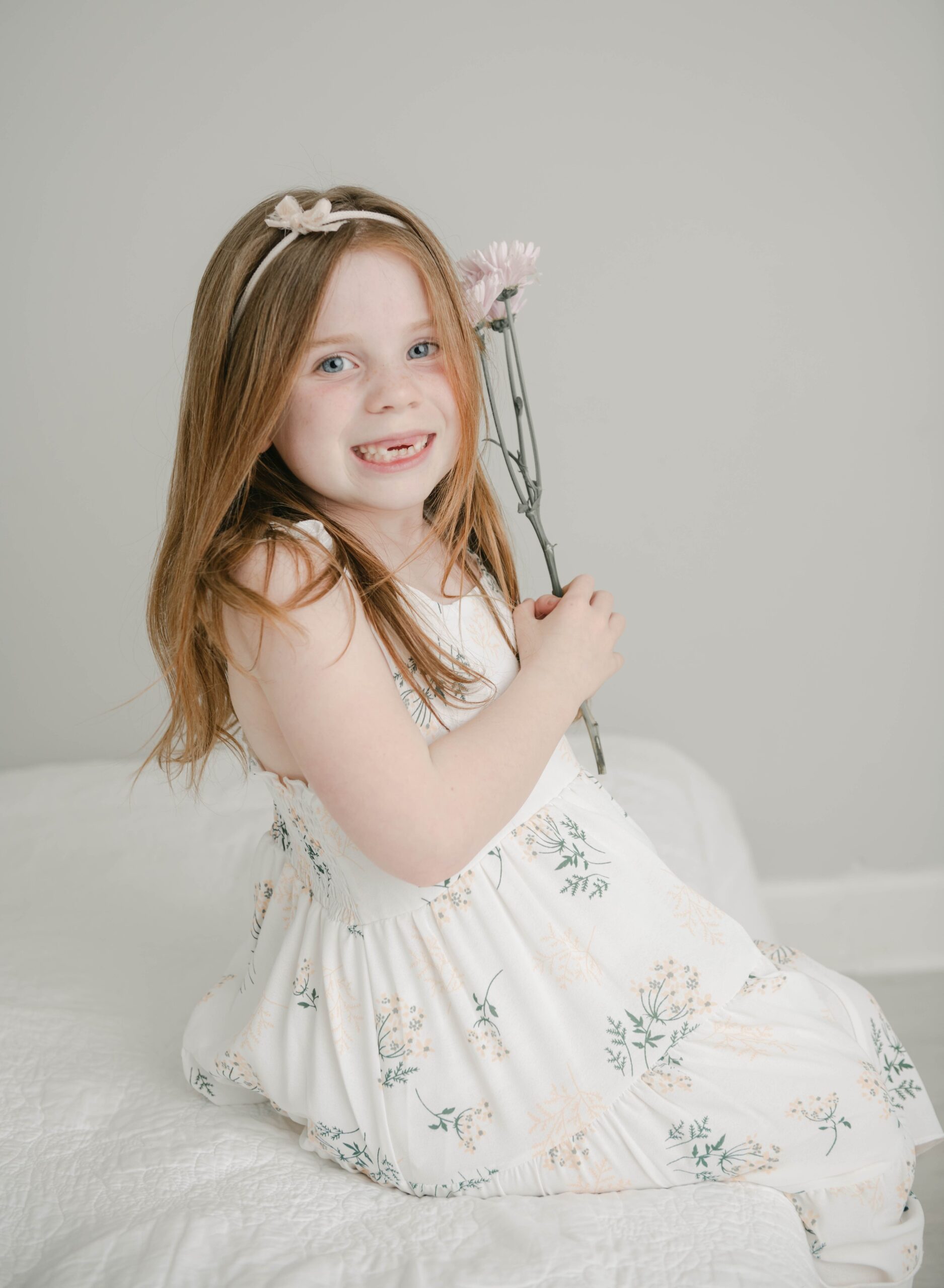 A young girl missing her two front teeth sits on the edge of a beed in a white dress holding some flowers budha babe