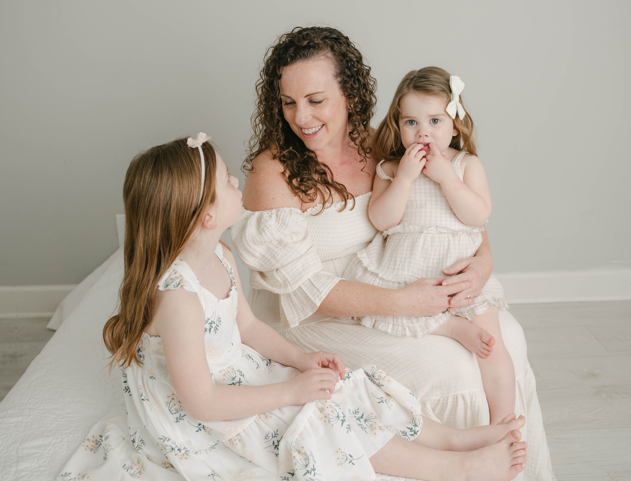 A mom sits on a small bed with her toddler daughter on her lap and older daughter next to her
