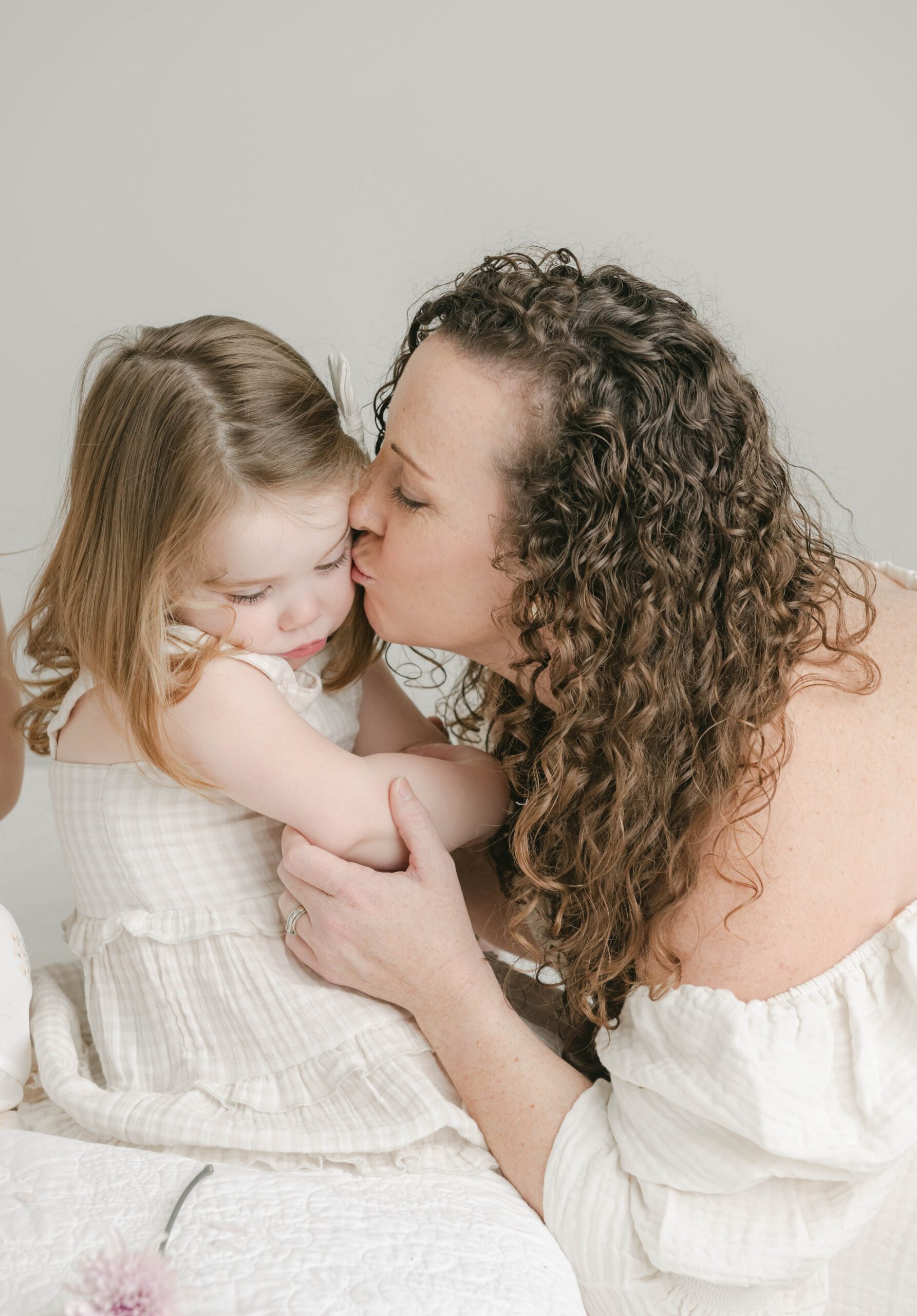 A mother kisses the cheek of her young daughter both in matching white dresses budha babe