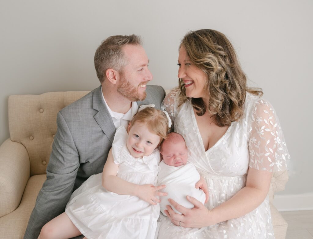 A mother and father sit on a tan couch in a studio with their newborn baby and toddler daughter