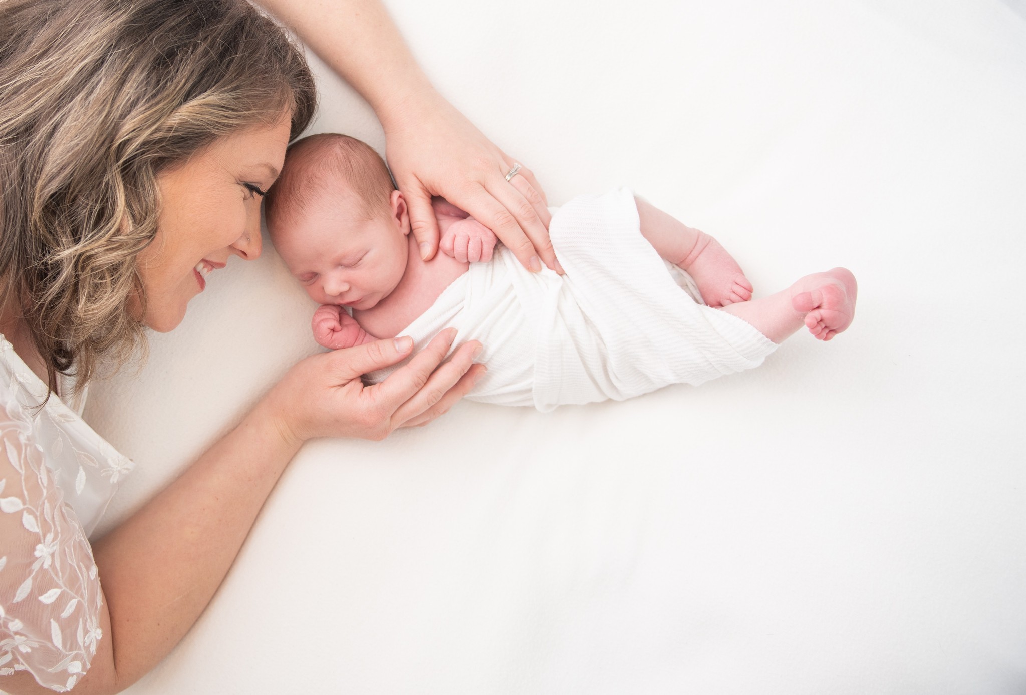 A newborn baby sleeps on a white bed with mom reaching over with cuddles