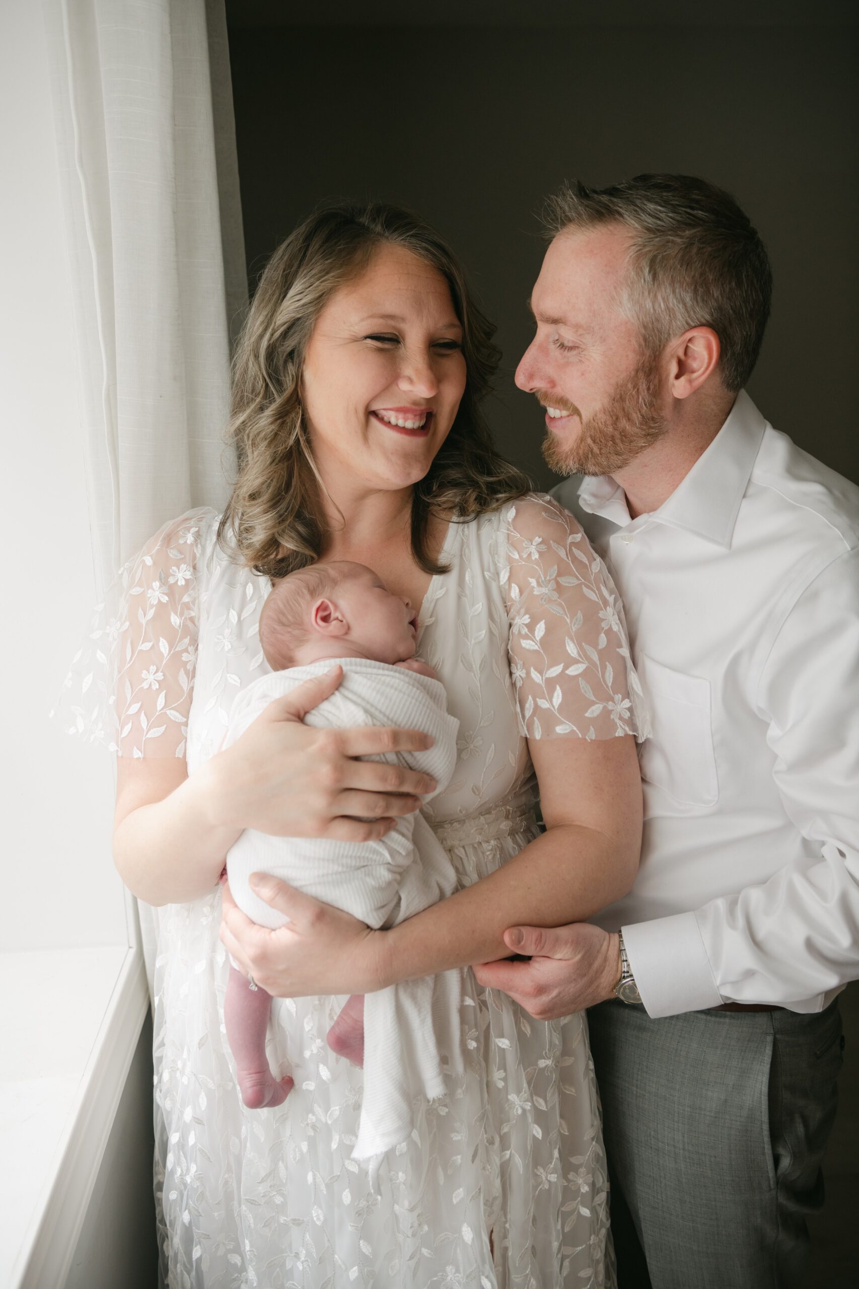 A mother holds her sleeping newborn baby against her chest in front of a window with dad leaning in rising moon midwifery