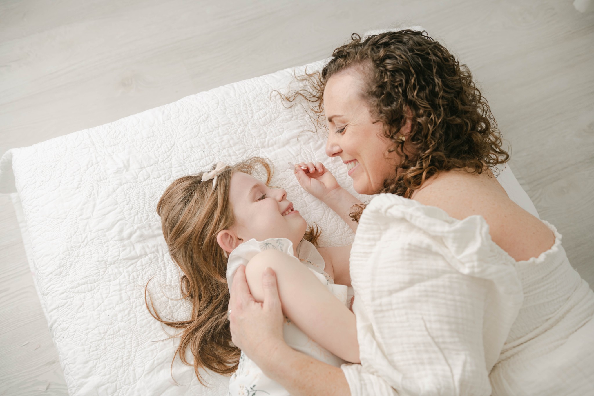 A mother lays on a blanket cuddling and playing with her young daughter in white dresses