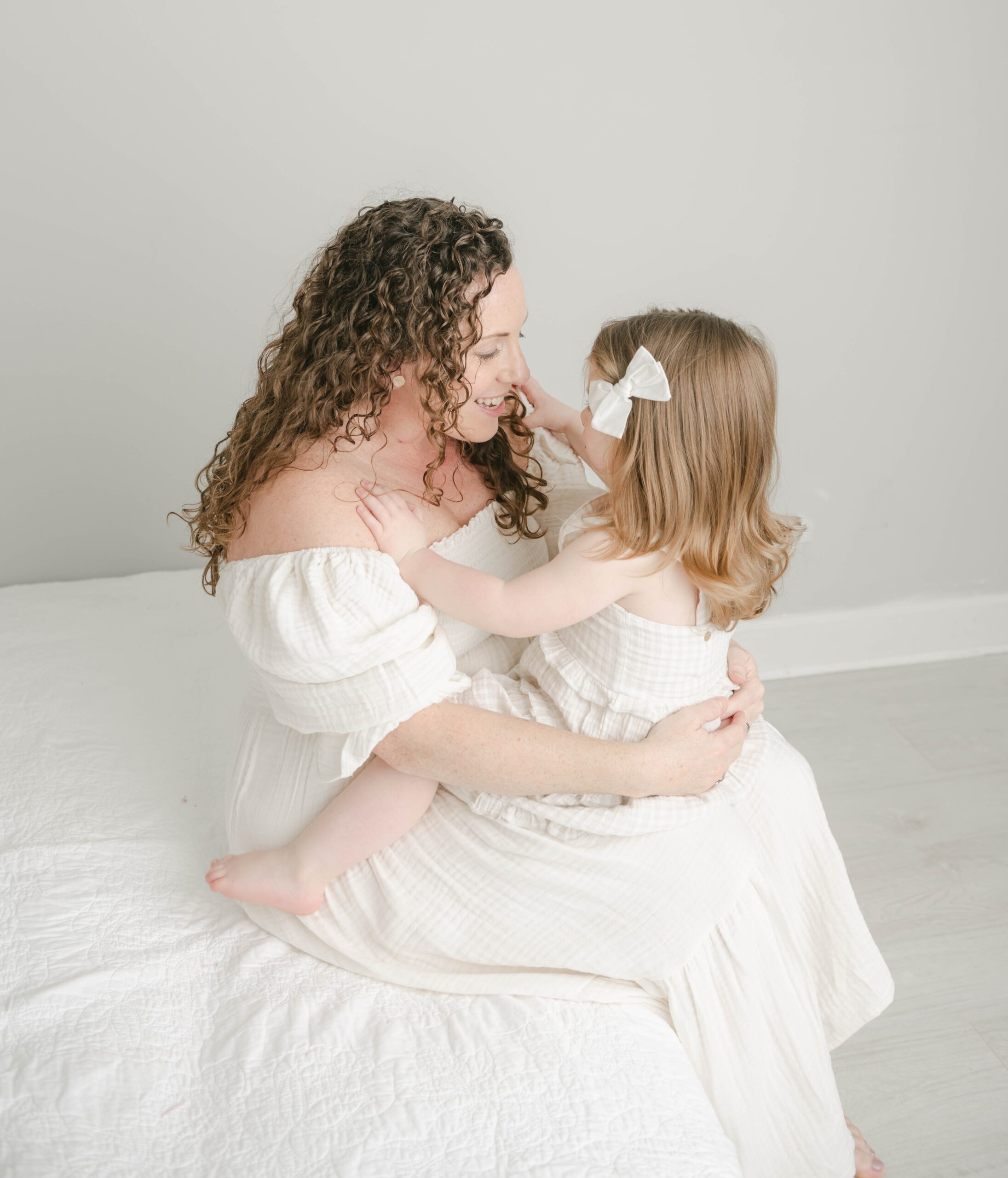 A mother sits on the edge of a bed playing with her young daughter in her lap in a studio Chester county obgyn