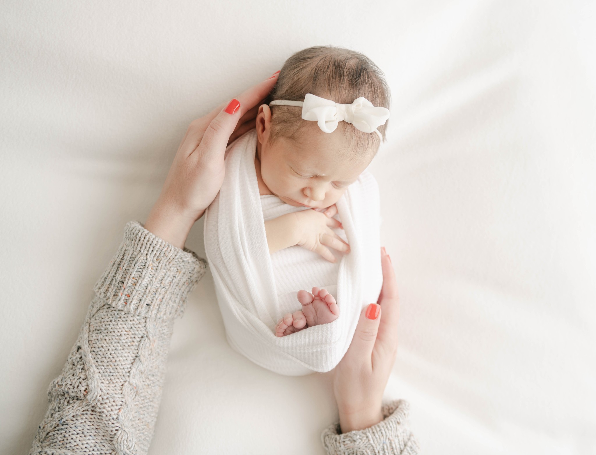 A mother places her hands on her sleeping newborn baby in a white swaddle
