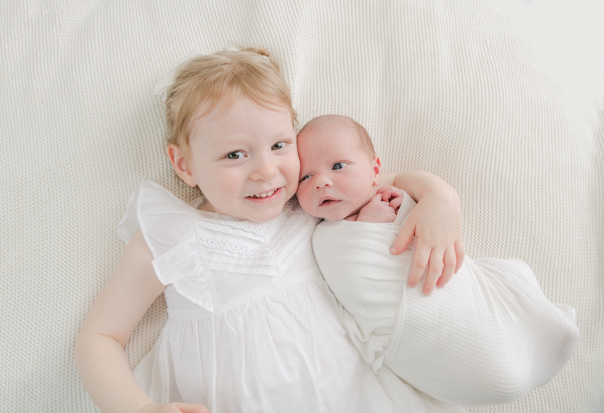 A young girl in a white dress lays on a bed cuddling with her newborn baby sibling chester county pediatricians