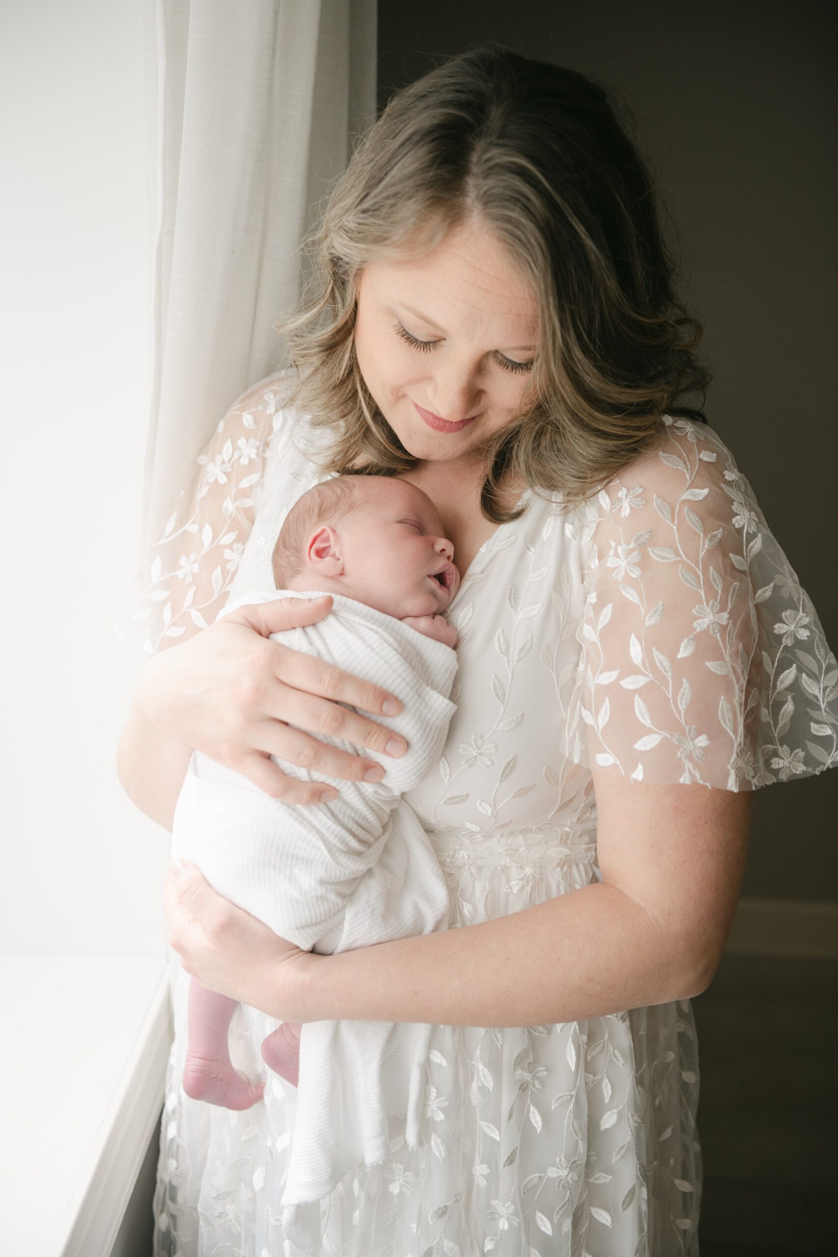 A mother stands in a window cradling her sleeping newborn on her chest prenatal massage lancaster pa