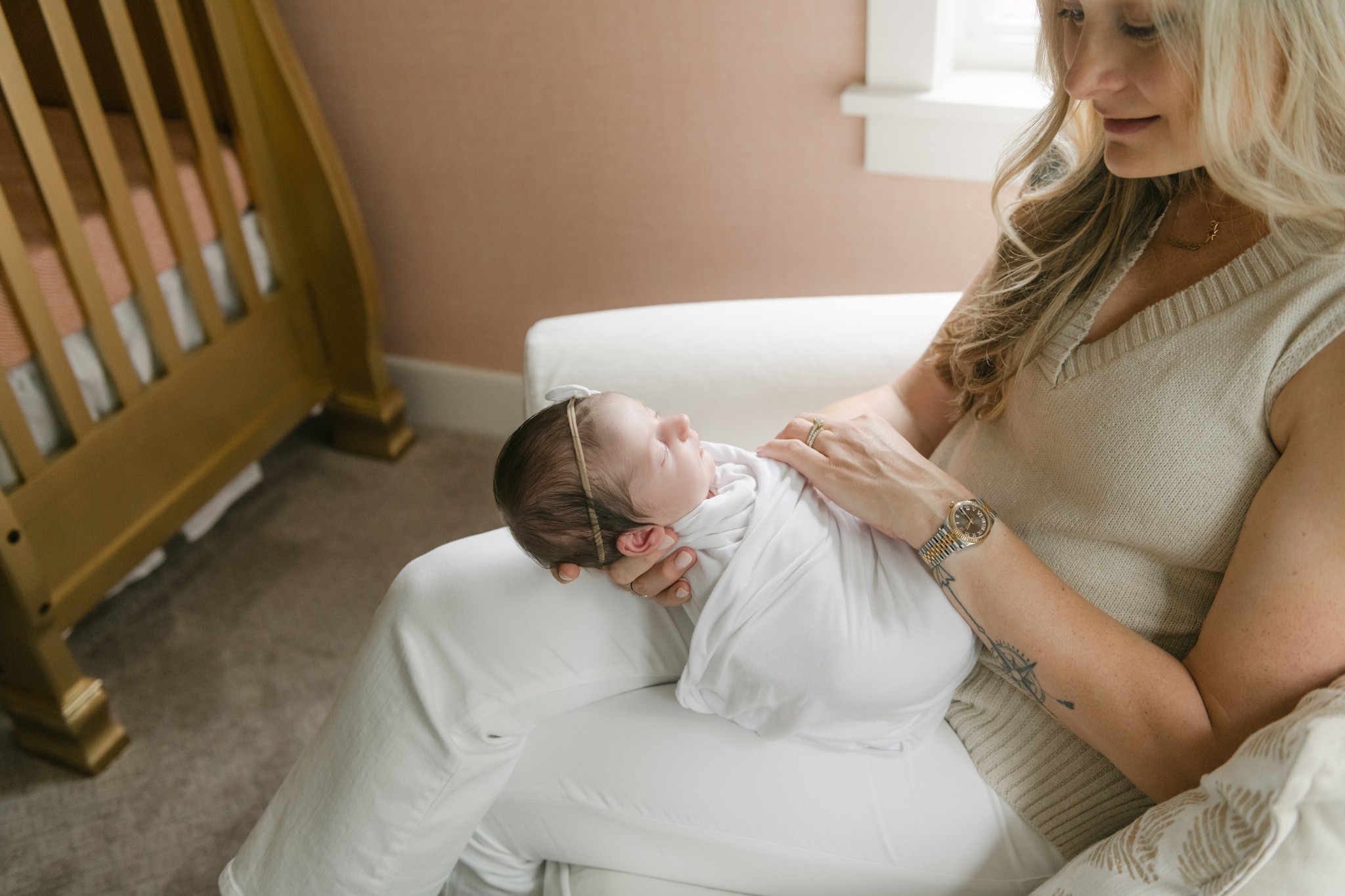 A newborn baby sleeps in a white swaddle while being held in mom's lap in a nursery