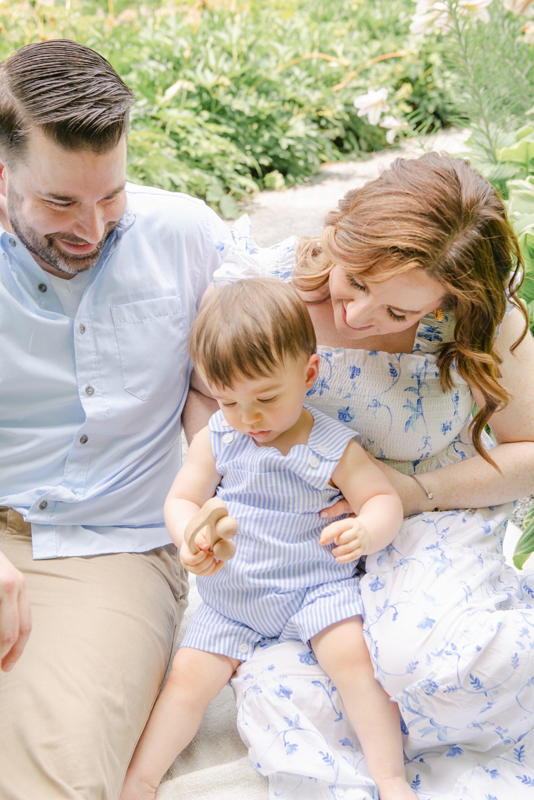 A mother and father sit on a picnic blanket in a garden path playing with their infant son with a wooden toy downington preschools