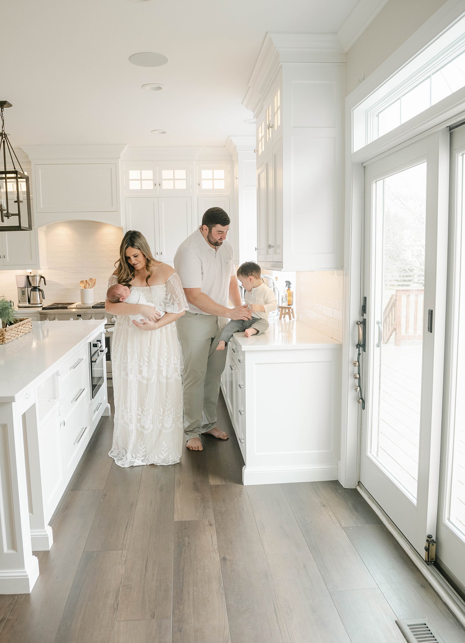 A mother and father play with their newborn baby and toddler son in the kitchen on the counter