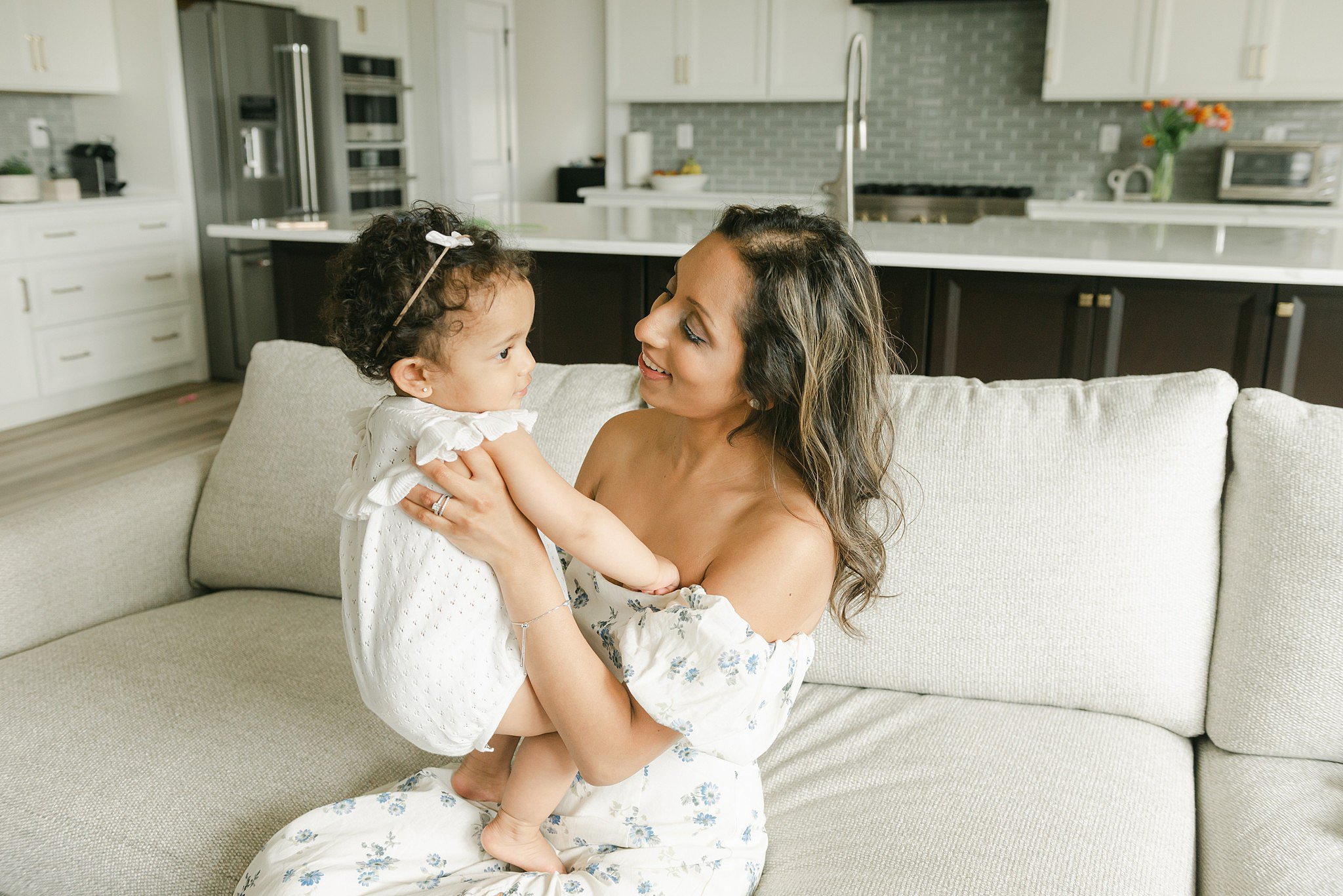 A mom in a white dress sits on a couch playing with her infant daughter in a white onesie downington daycare