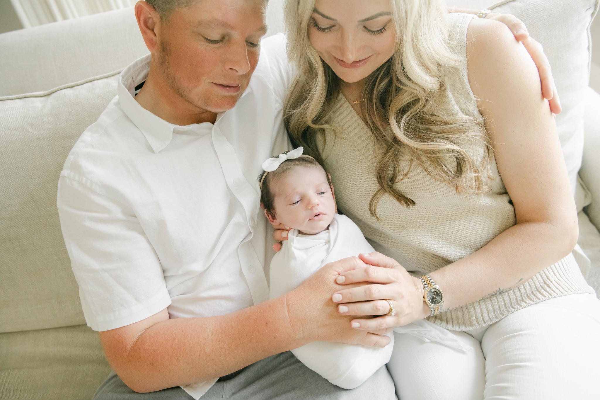 A mom and dad sit on a couch with their newborn daughter in their laps between them under a window after going to lifecycle womancare
