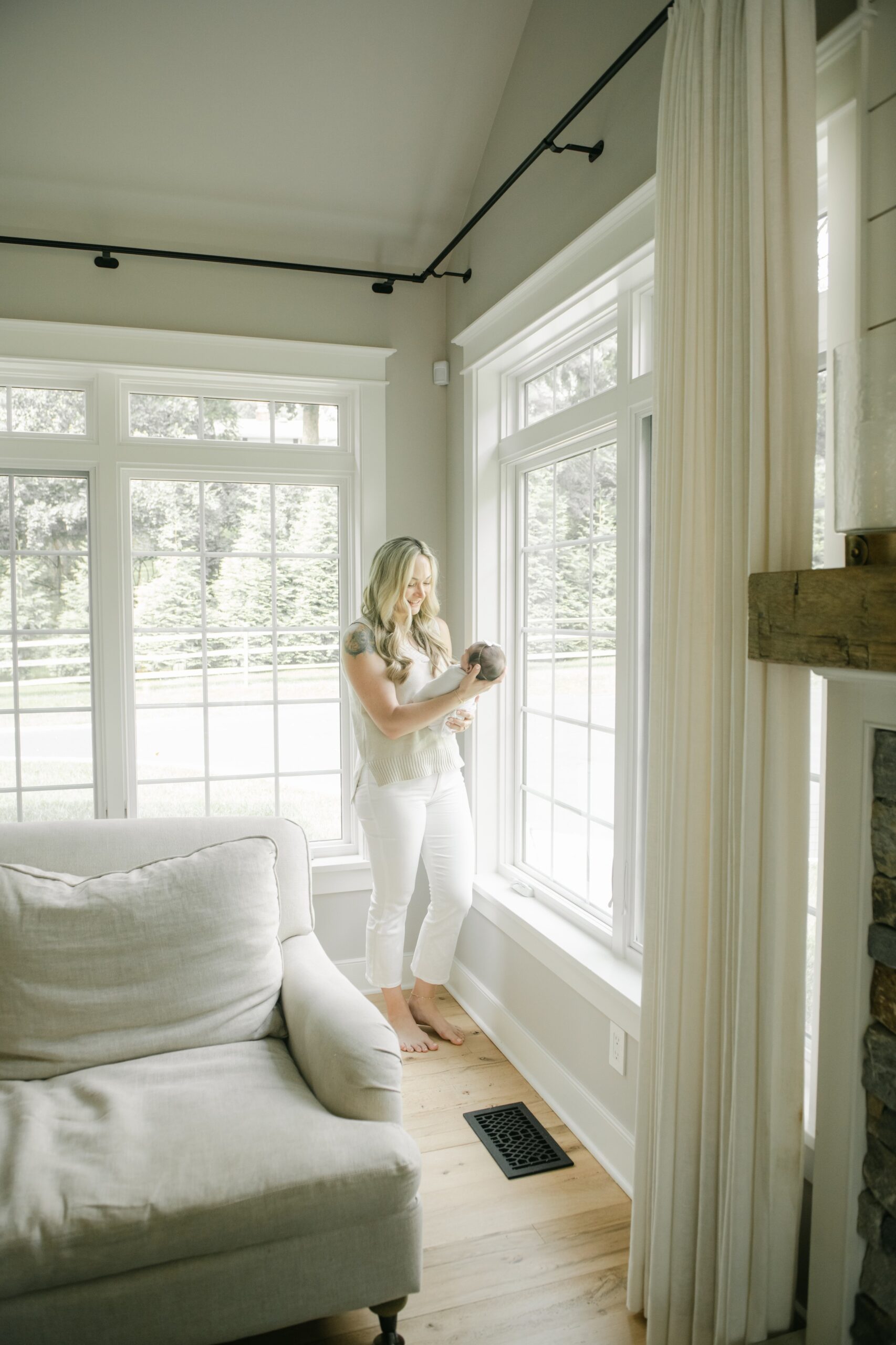 A new mother in white pants and green shirt stands in a large window smiling down at her newborn baby in her hands after a visit to lifecycle womancare