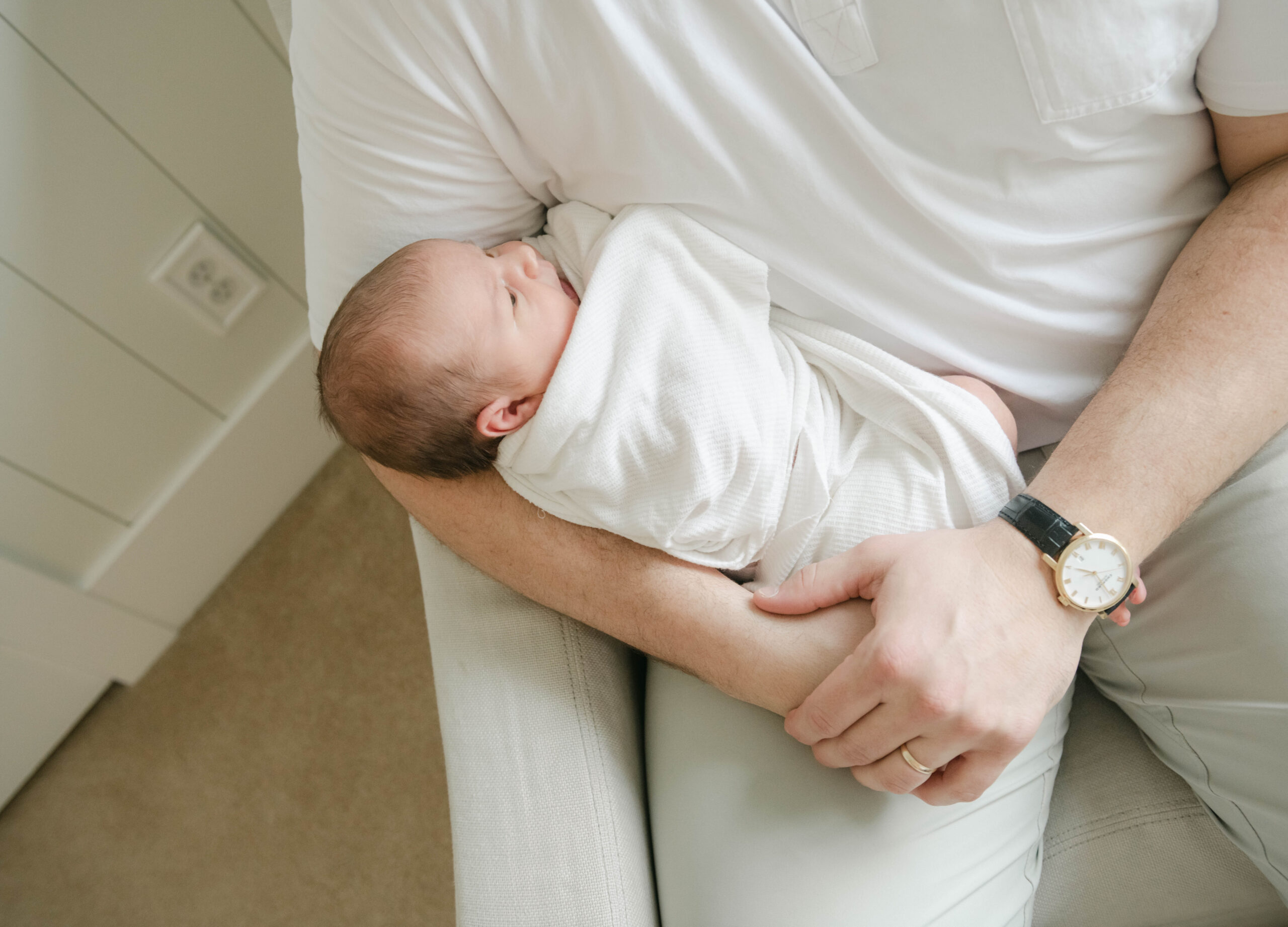 A newborn baby lays in dad's lap while wrapped in a white blanket as he sits in a nursery chair