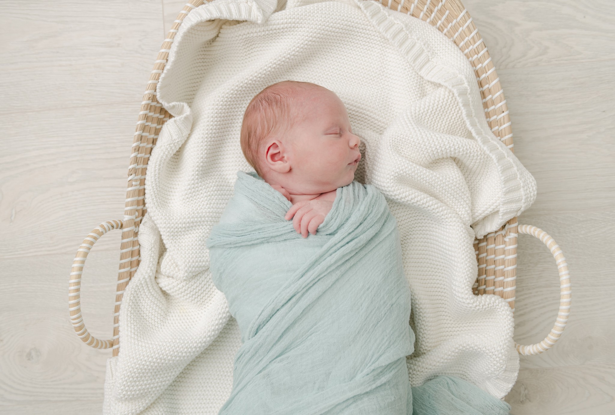A newborn baby sleeps in a woven basket and green swaddle