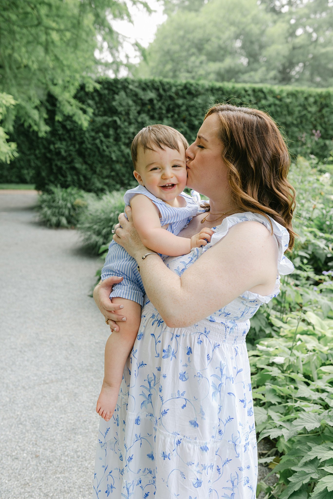 A mother in a blue dress stands in a garden kissing her toddler son in a blue onesie with the help of a nanny lancaster pa