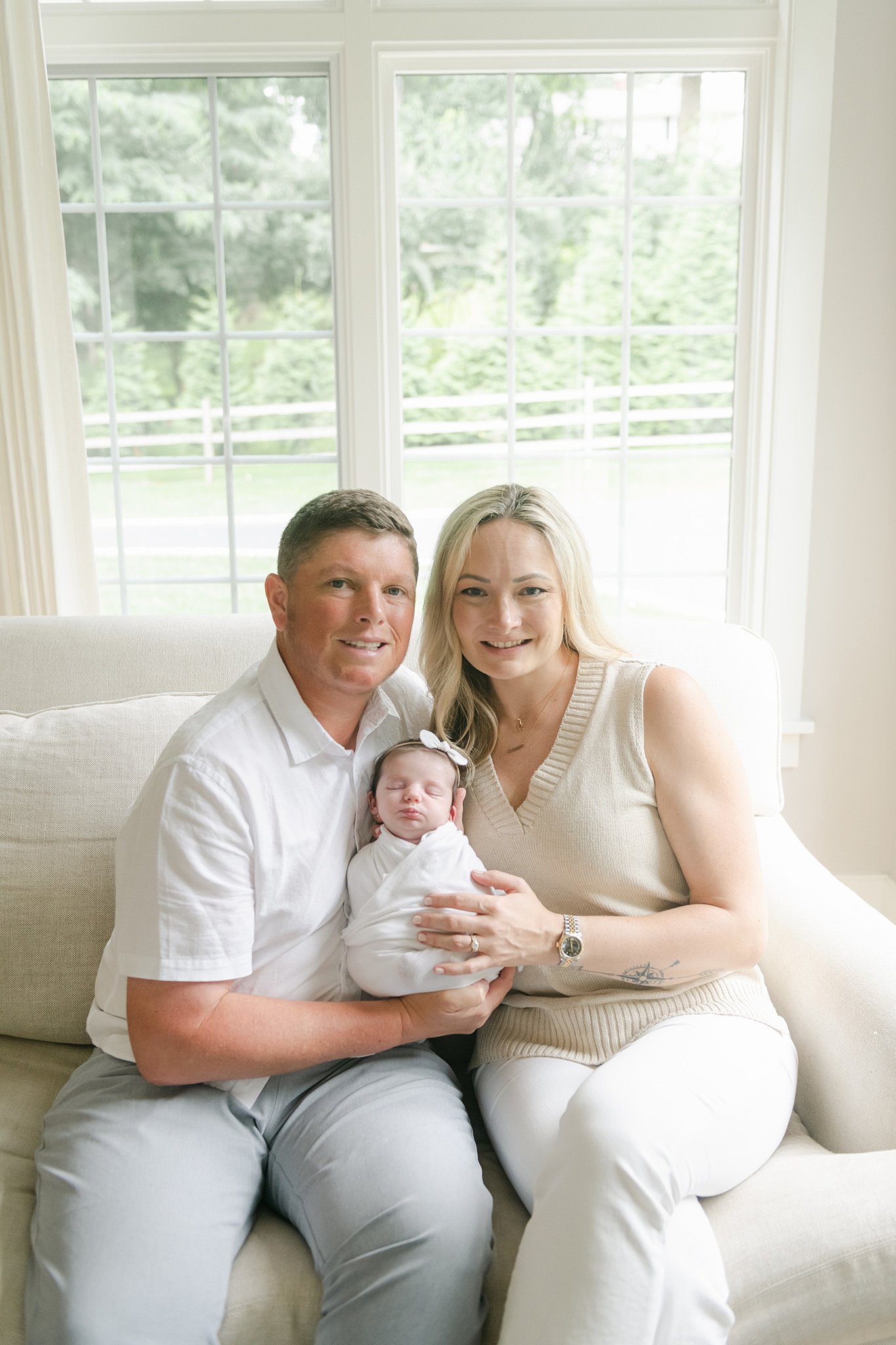 New parents sit on a couch by a window while holding their sleeping newborn daughter in a white swaddle after using prenatal yoga lancaster pa