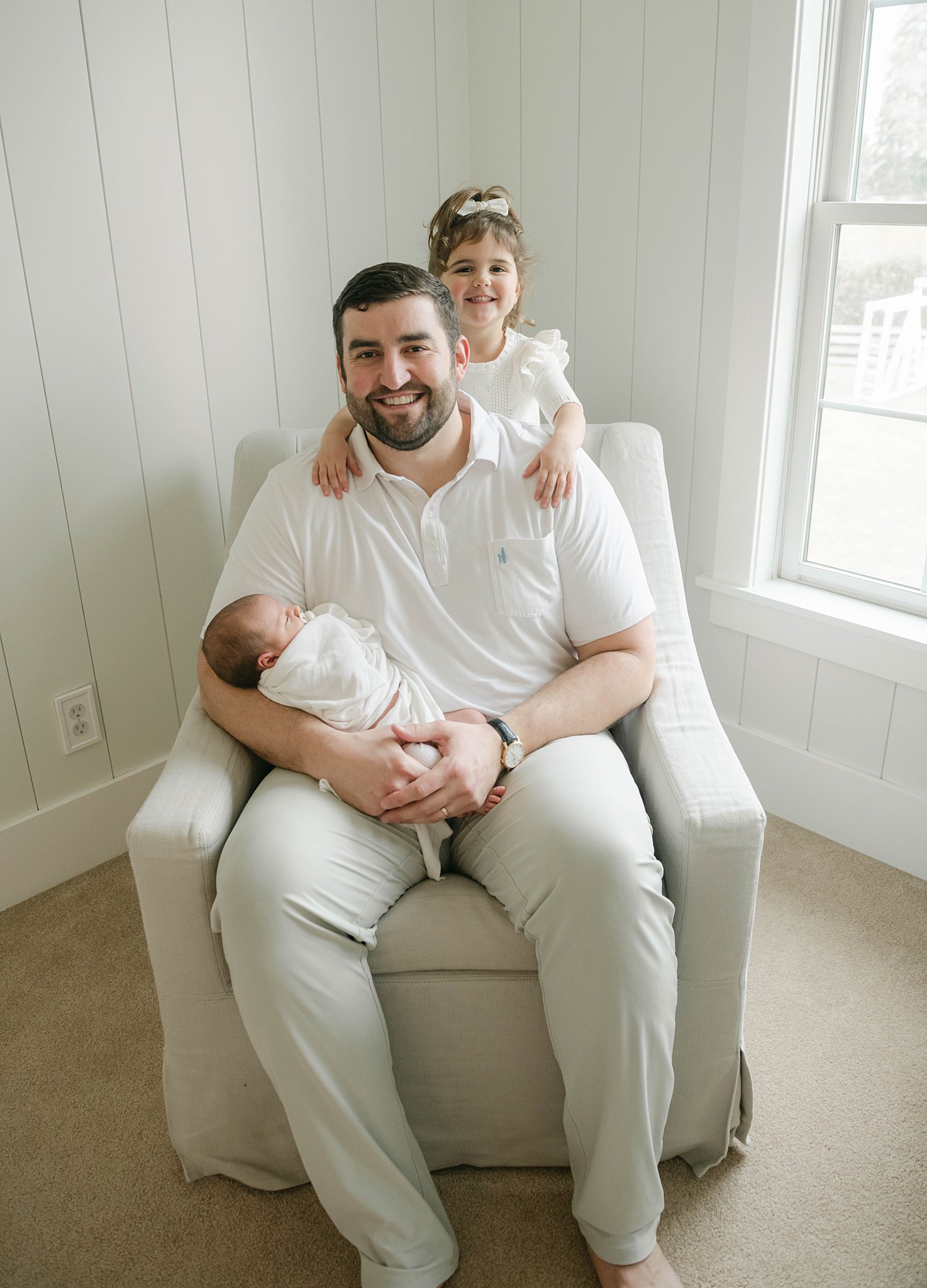 dad sits in an armchair wearing a white polo shirt with his daughter in a white dress and newborn