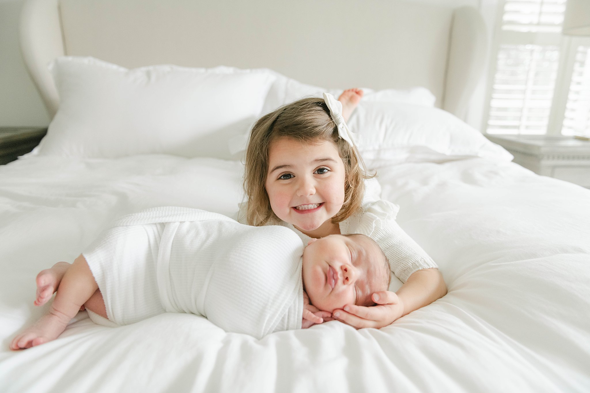 big sister holds the head of her sleeping newborn sibling on a white bed
