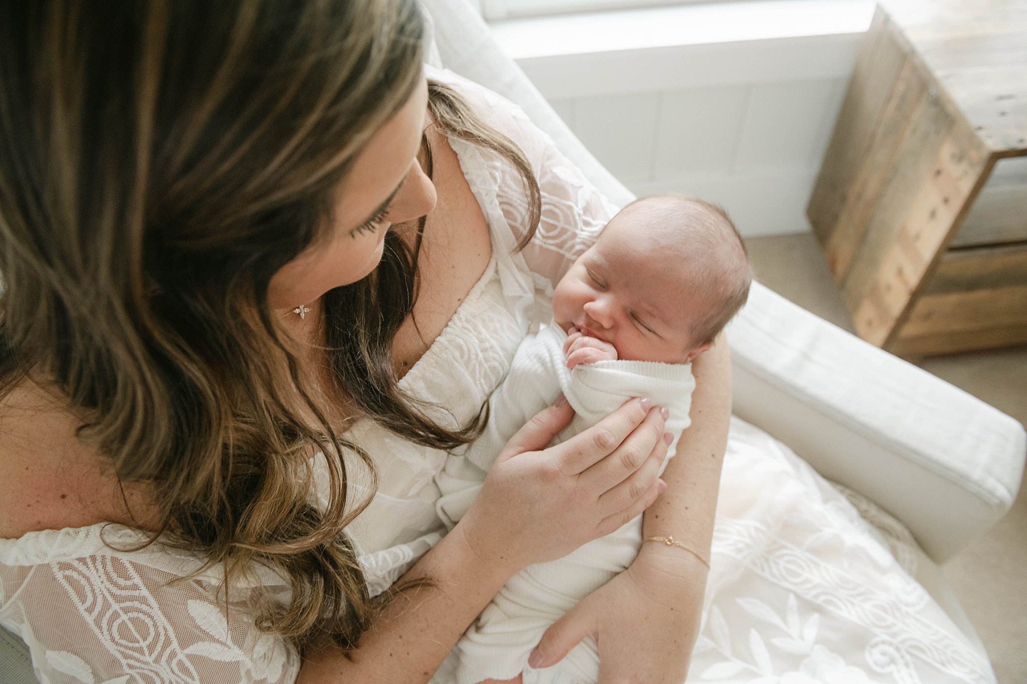 Mother sits in an armchair wearing white dress holding her sleeping newborn midwives lancaster