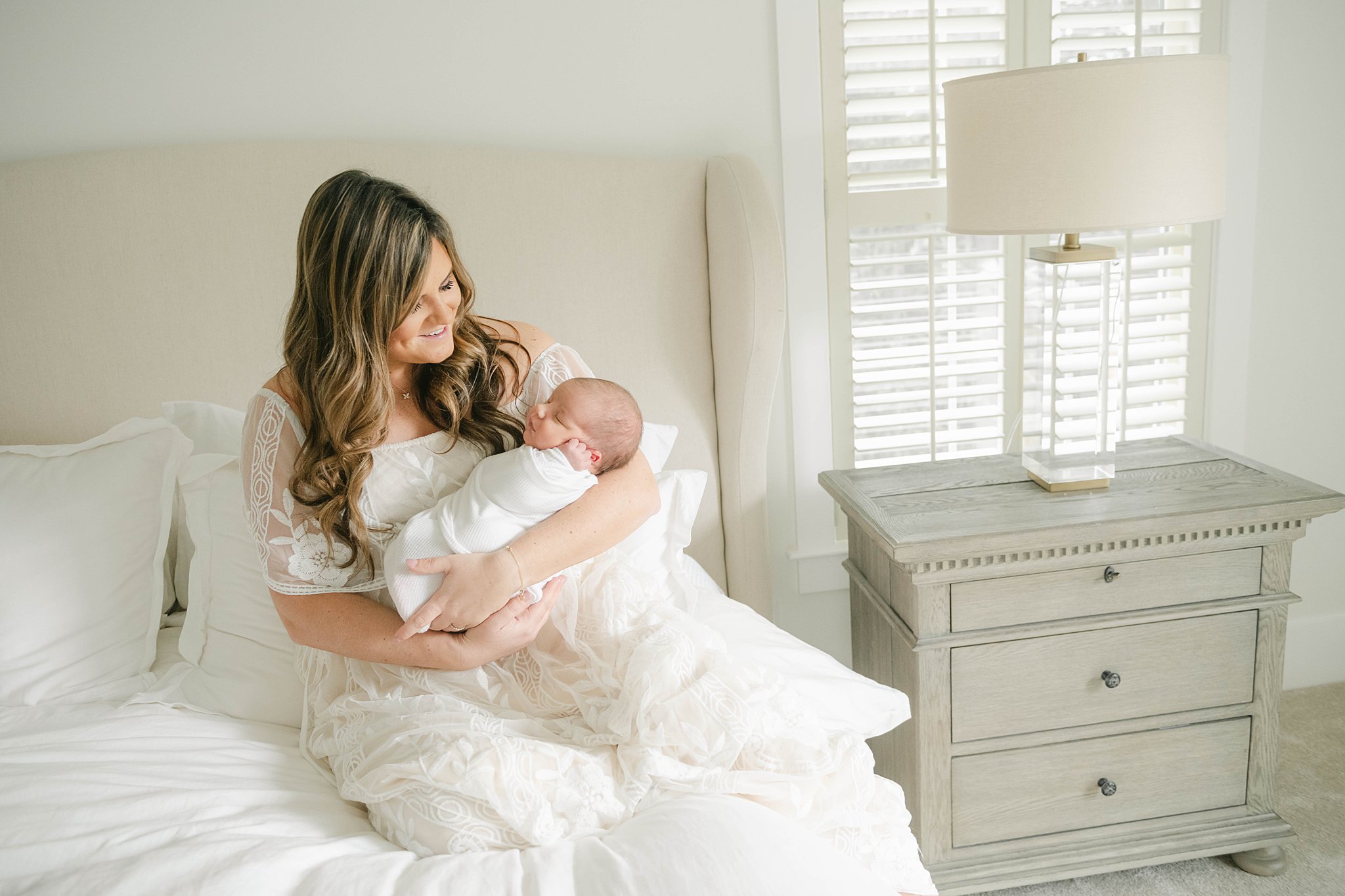 Mother in a white lace dress sits on a bed while holding her sleeping newborn