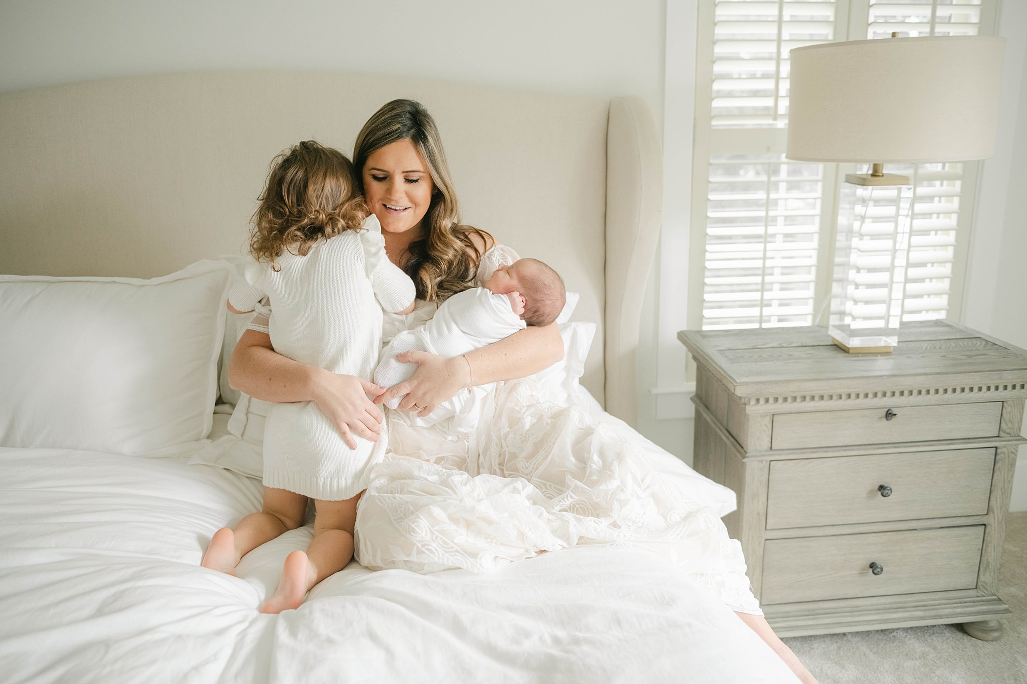 Mother in a white dress gets hugged by her toddler while holding her newborn on a bed Labor of Love Doula Services