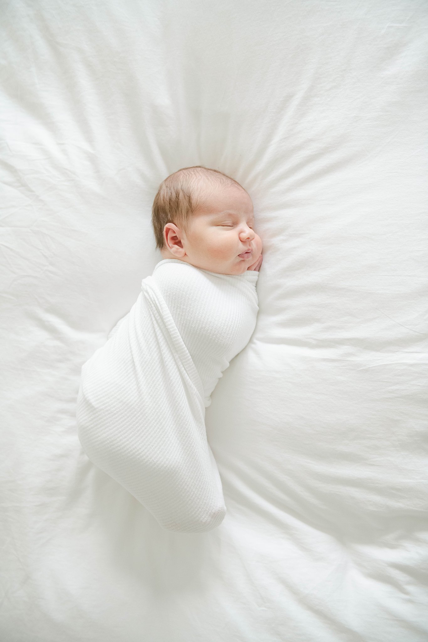 Newborn baby sleeps swaddled in a white blanket on a white bed with hands under head Labor of Love Doula Services
