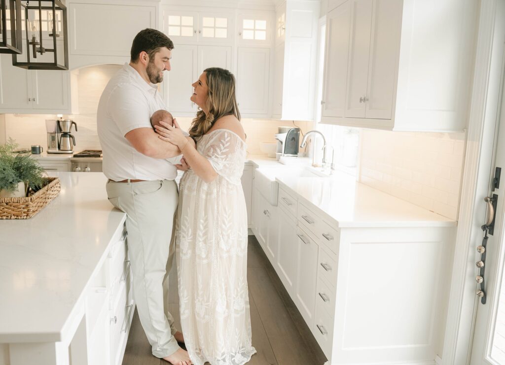mom and dad stand in a white kitchen while holding their newborn child