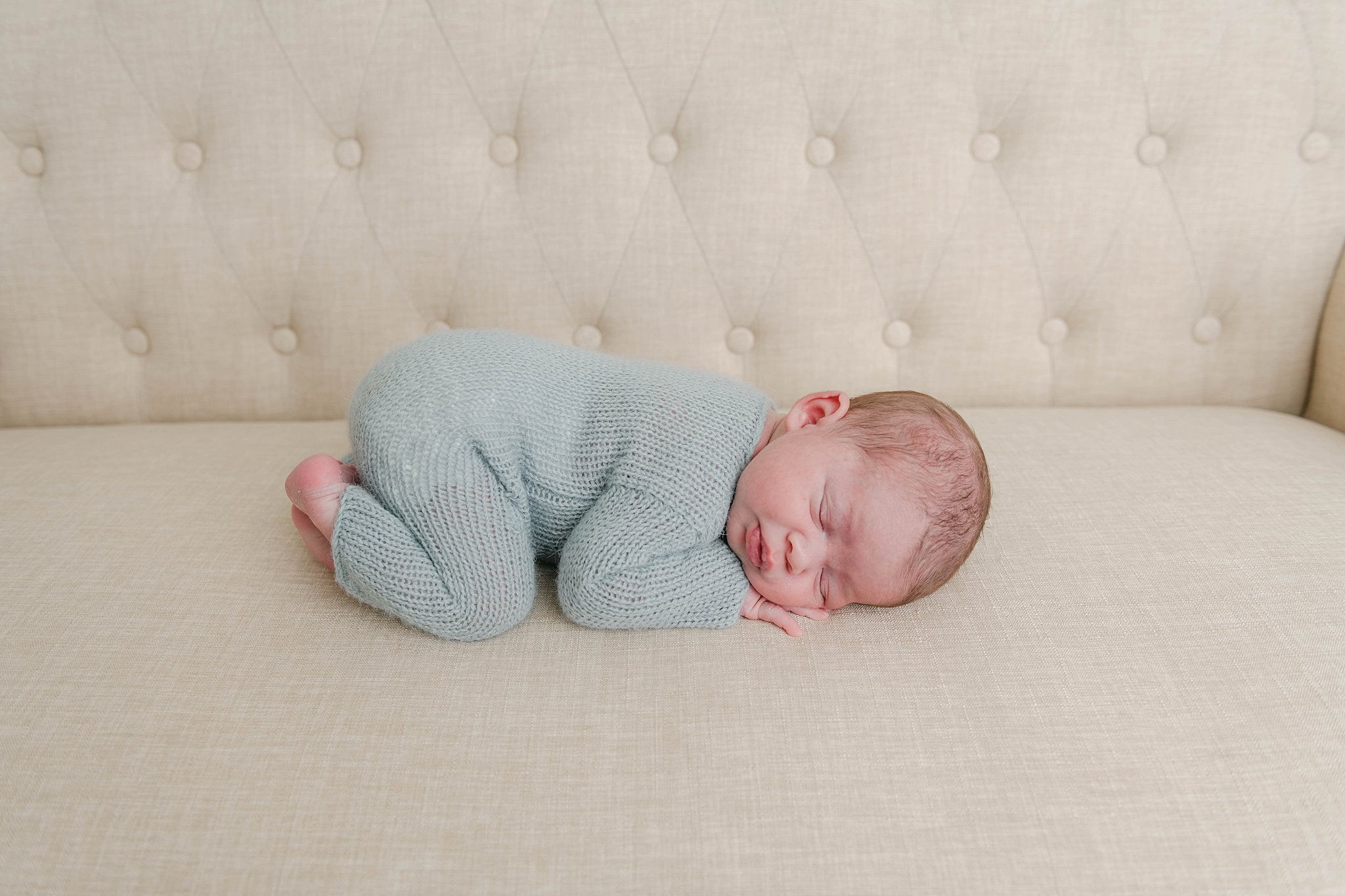 A newborn baby sleeps in a blue knit onesie on its knees and hands on a tan couch before we meet