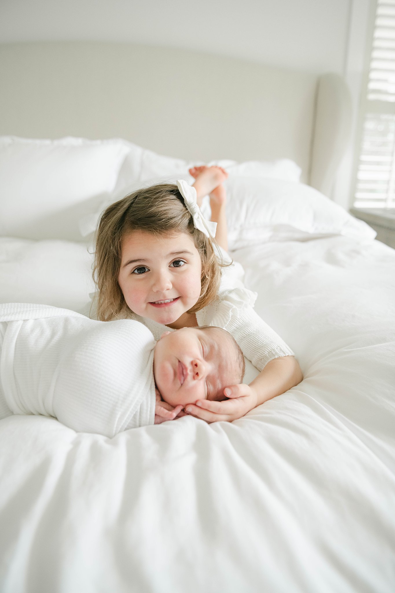 A young girl lays on a bed while holding the head of her sleeping newborn sibling