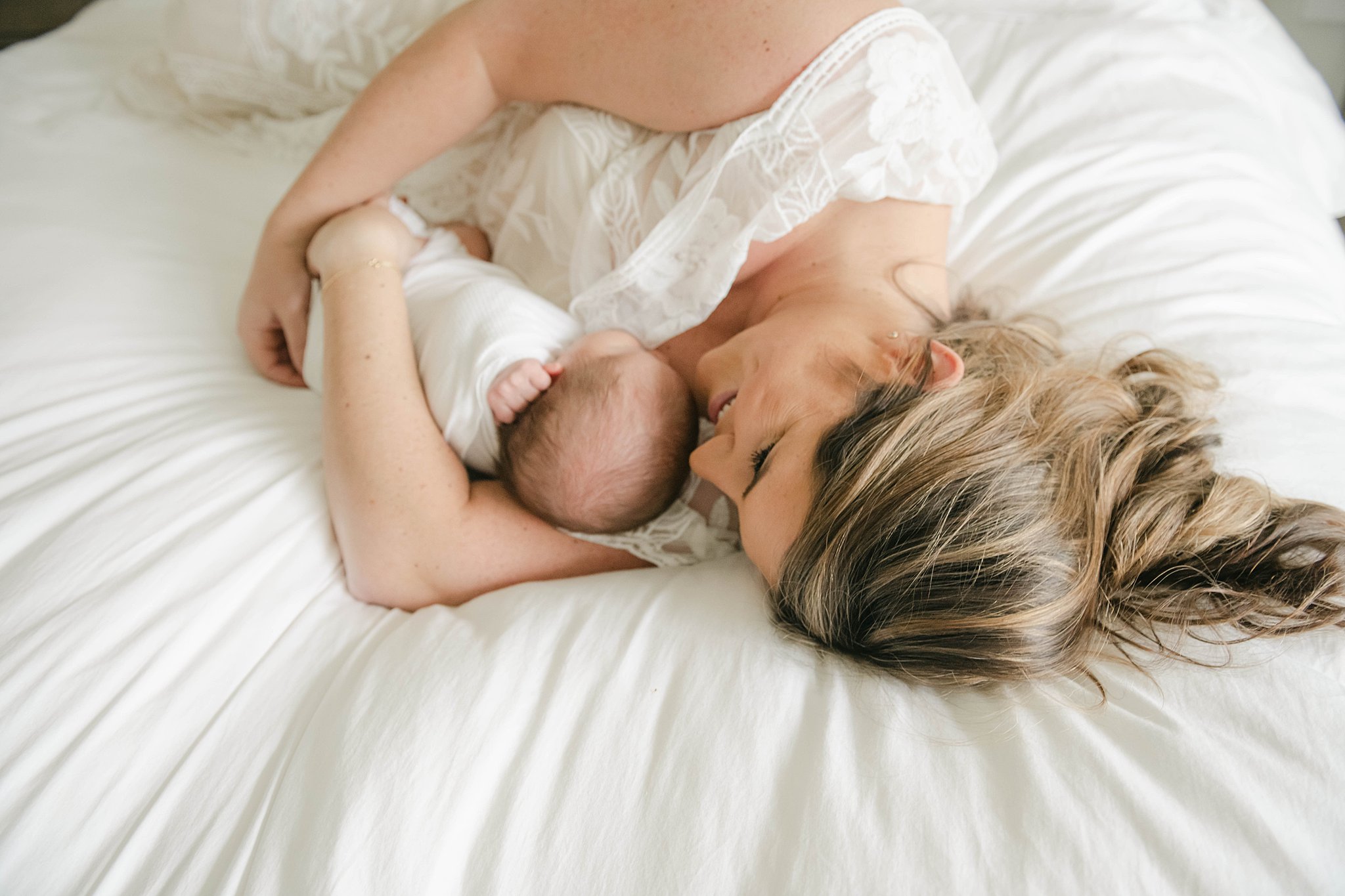 A mother cuddles on a bed with her newborn chiile while wearing a white dress blossoming bellies