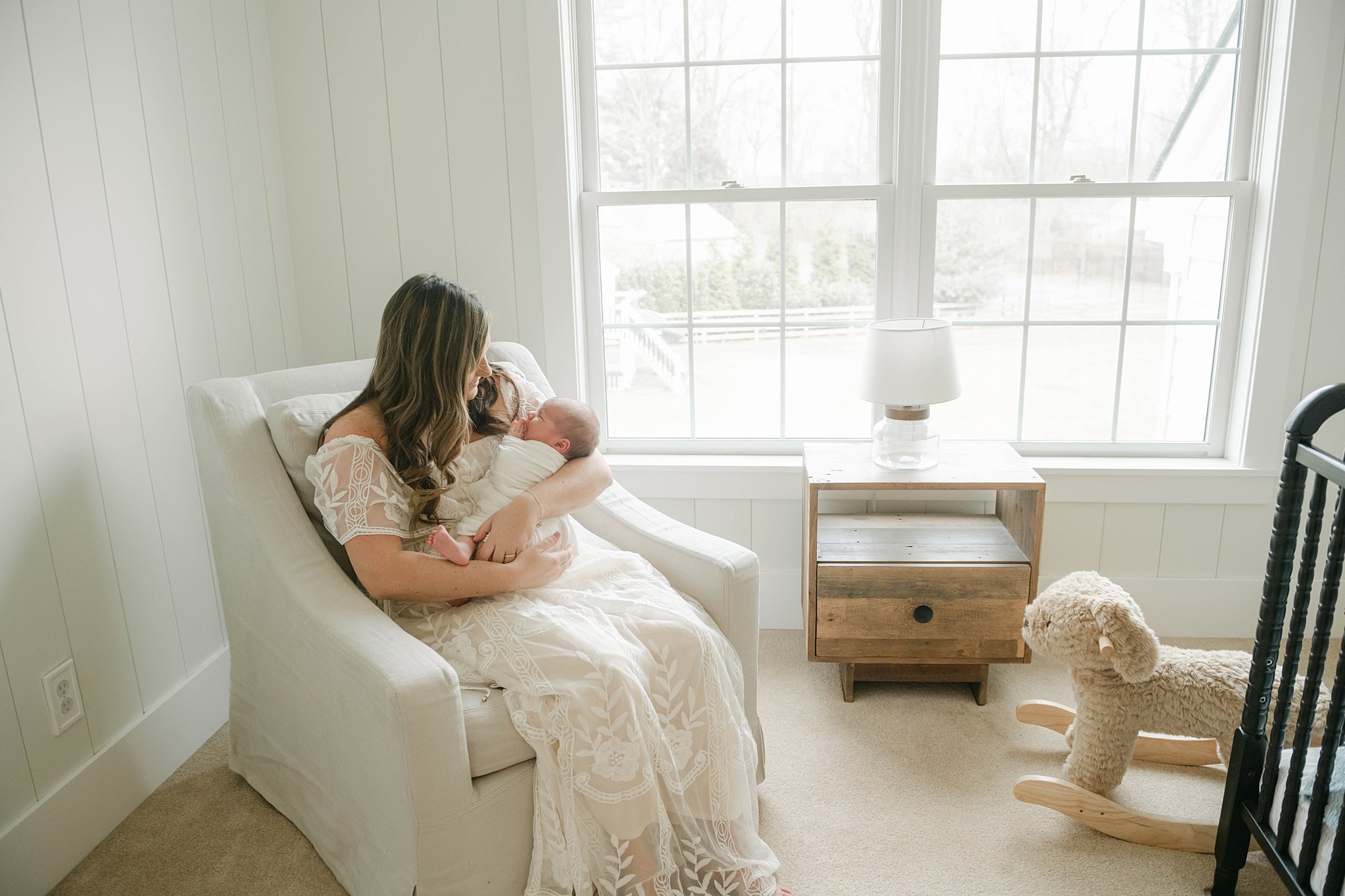 A mother sits in a nursery chair in front of a window with a soft rocking horse and rustic side table while holding her newborn baby blossoming bellies