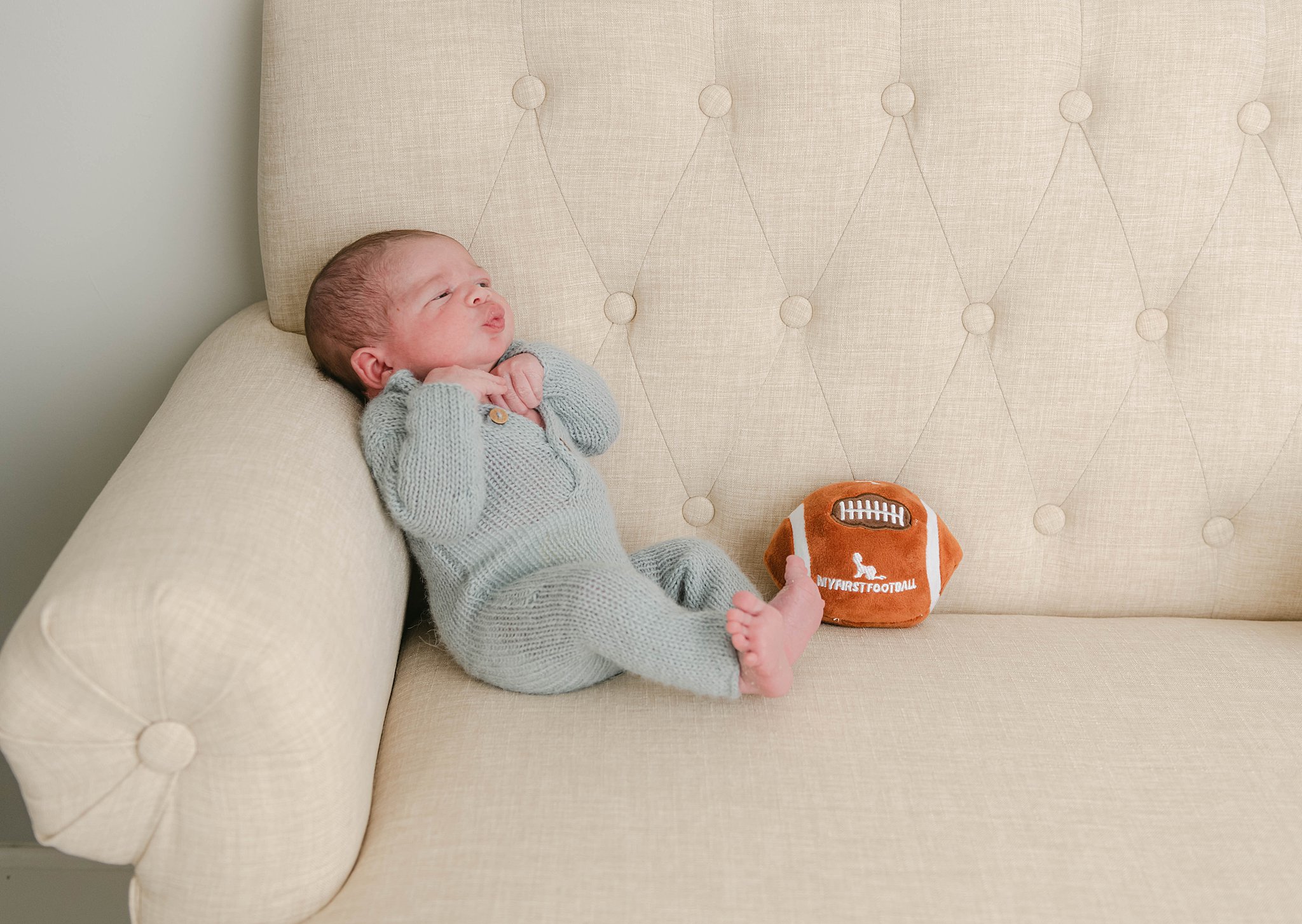 A newborn baby boy sits on a beige chair with a stuffed football toy while making a silly face the nesting house
