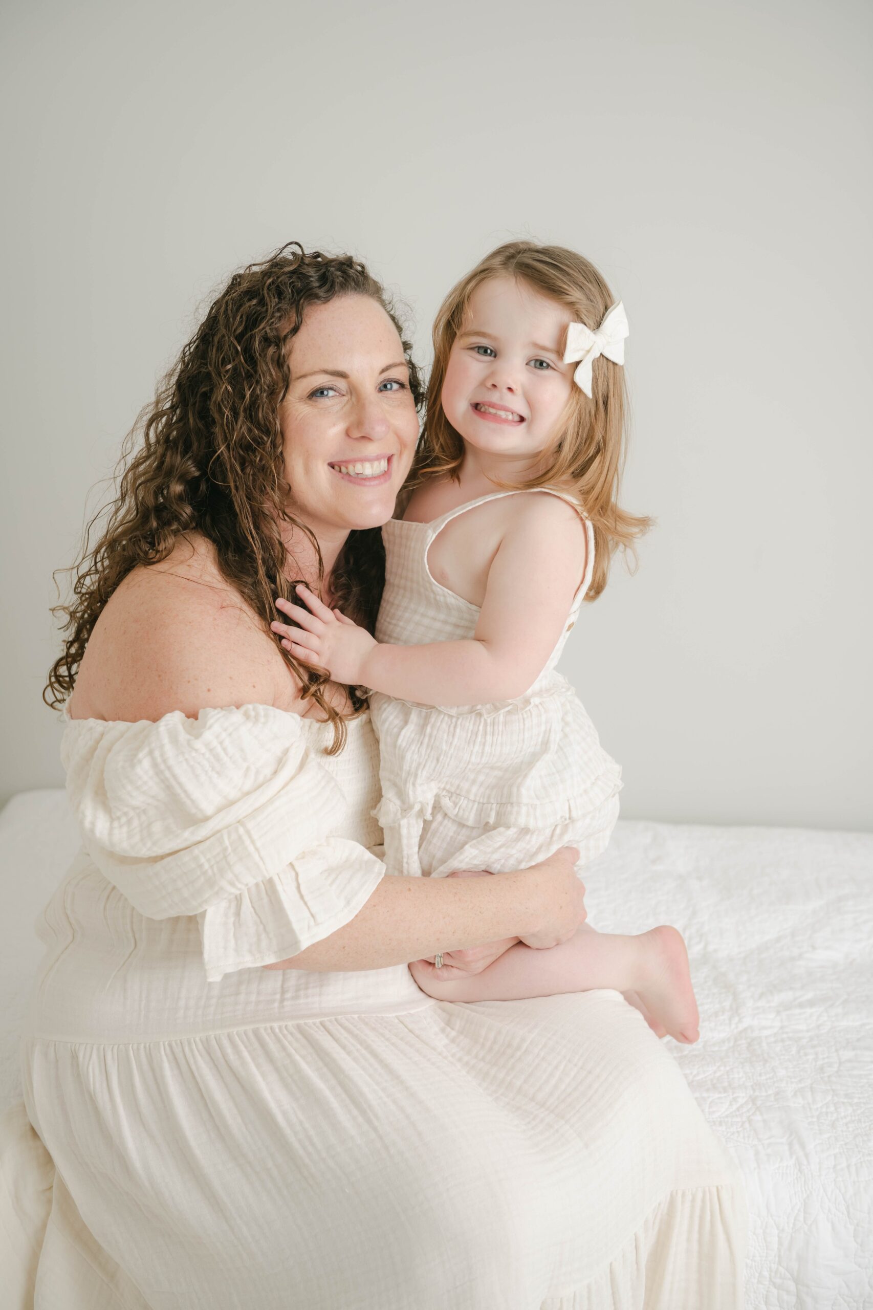 A mother sits on the edge of a white bed with her toddler daughter kneeling on her leg bellaboo lancaster