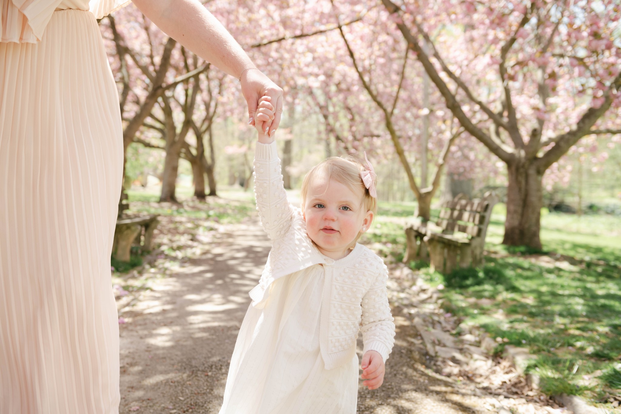 A young girl in a white dress and sweater walks while holding her mom's hand through a park h rose boutique