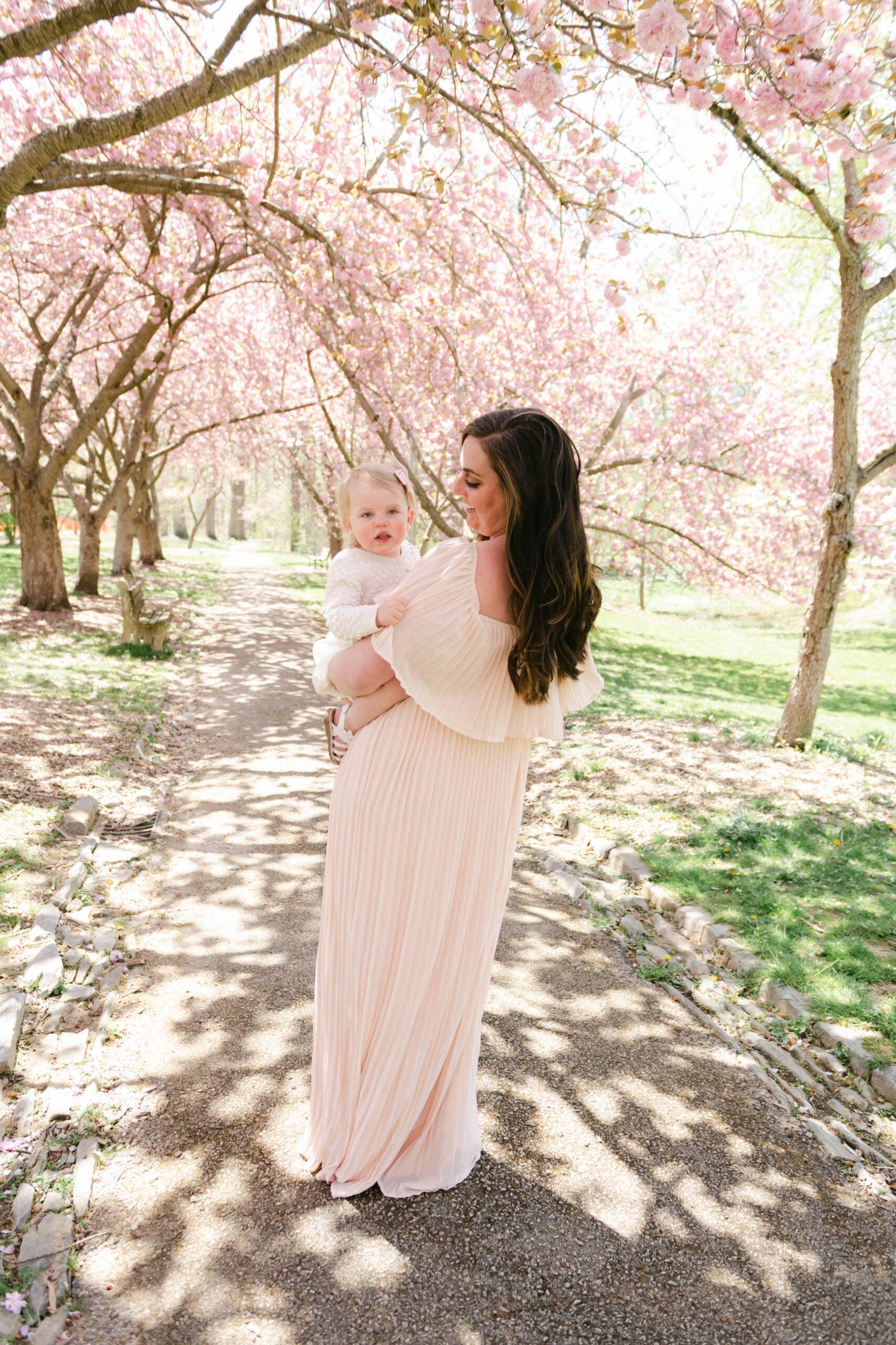 A mother in a beige dress holds her toddler daughter on her hip in a park path lined with pink dogwood trees