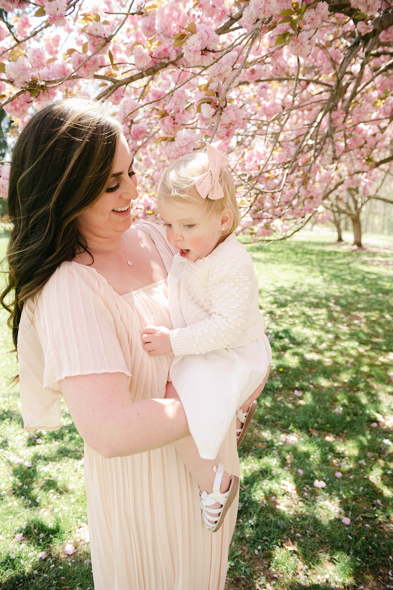 A mother holds her young daughter wearing a white dress and pink bow under pink blossoms of a tree kids 'n kribs