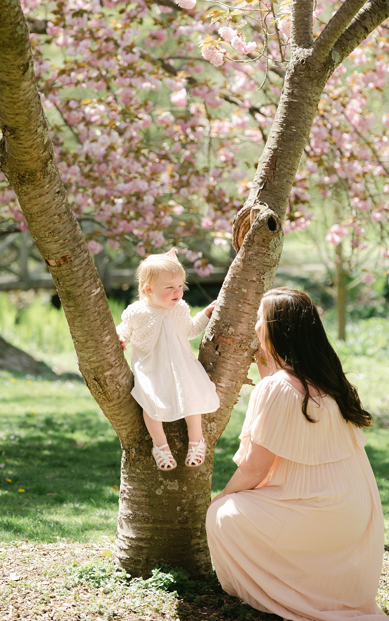 A young toddler sits in a tree in a white dress while mom kneels next to her kids 'n kribs