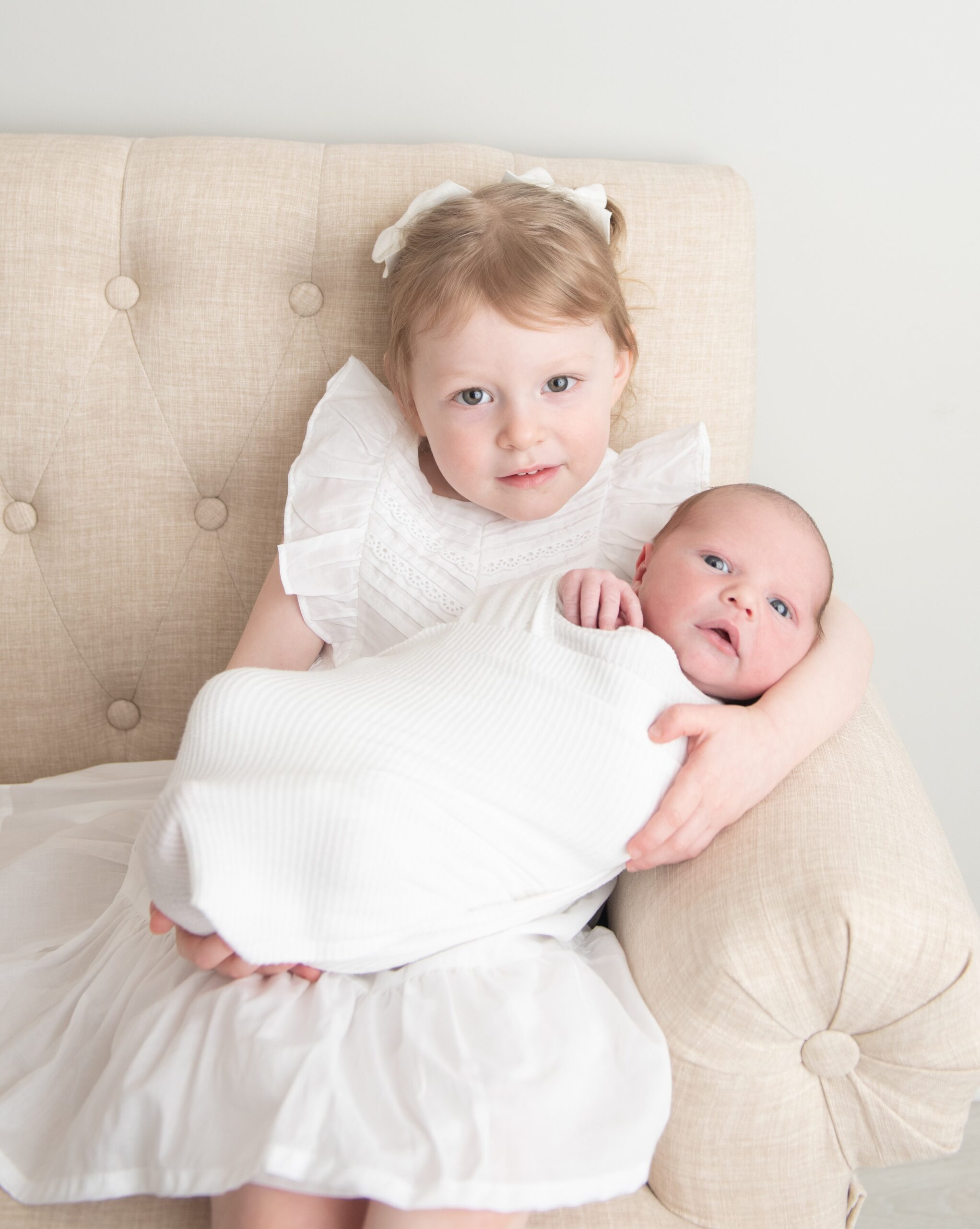 A young girl in a white dress sits on a couch with her newborn baby sibling in her lap