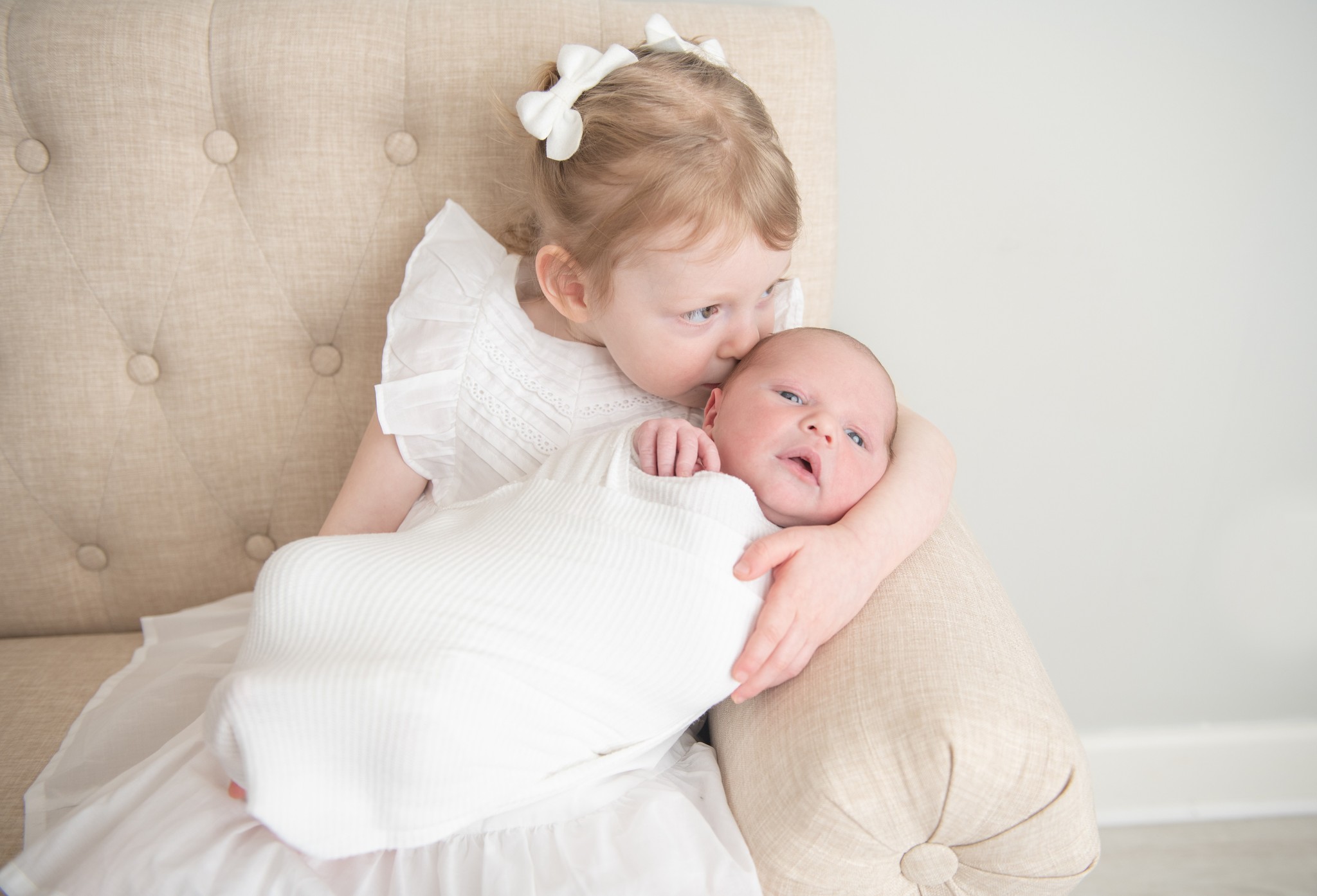 A young girl sits on a couch holding and kissing her newborn baby sibling the blue beret