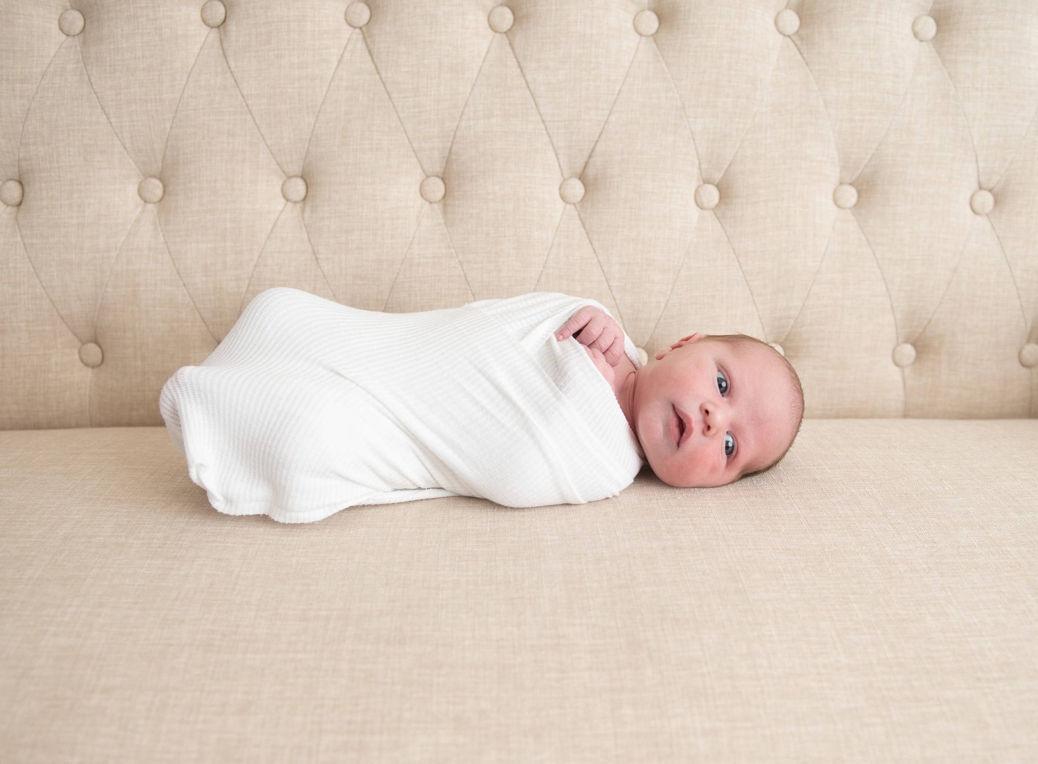 A newborn baby lays on a bench in a white swaddle with eyes open the blue beret