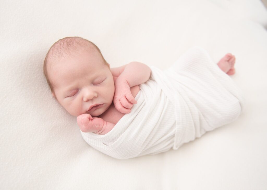 A newborn baby sleeps in a white swaddle with hands and feet exposed