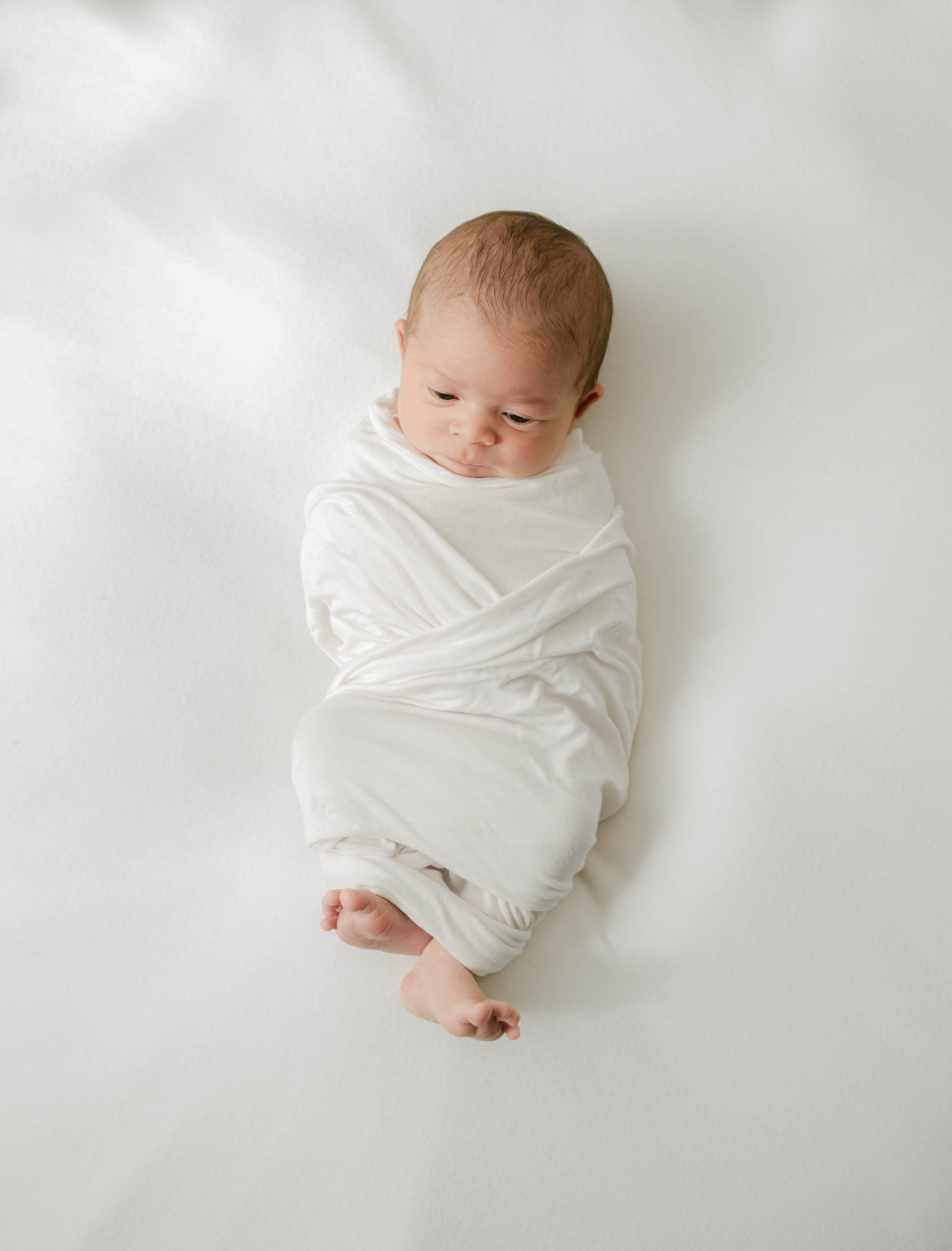 A newborn baby in a white swaddle looks at its feet on a white bed lactation consultant lancaster pa