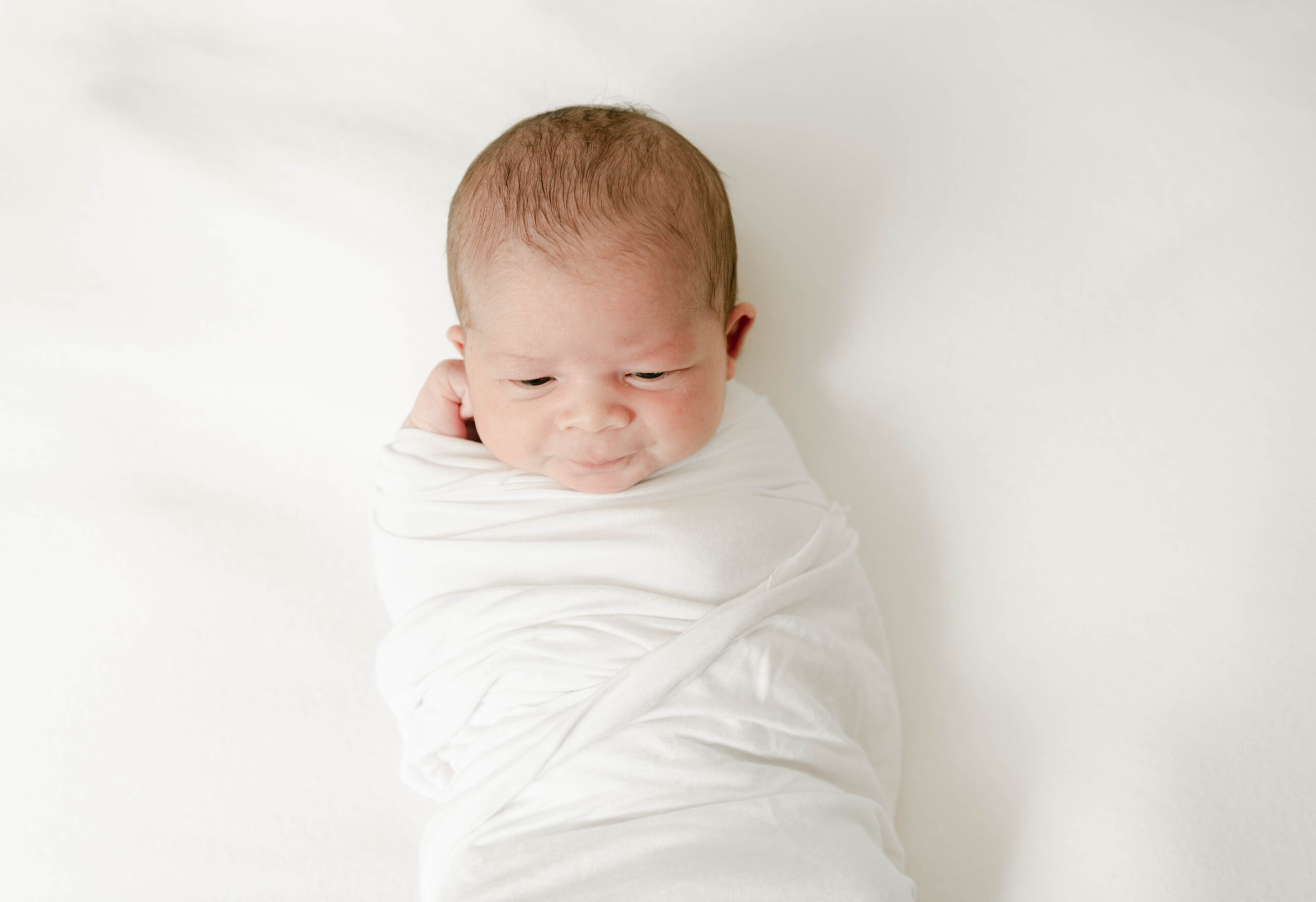 A newborn baby lays on a white bed in a white swaddle with eyes open