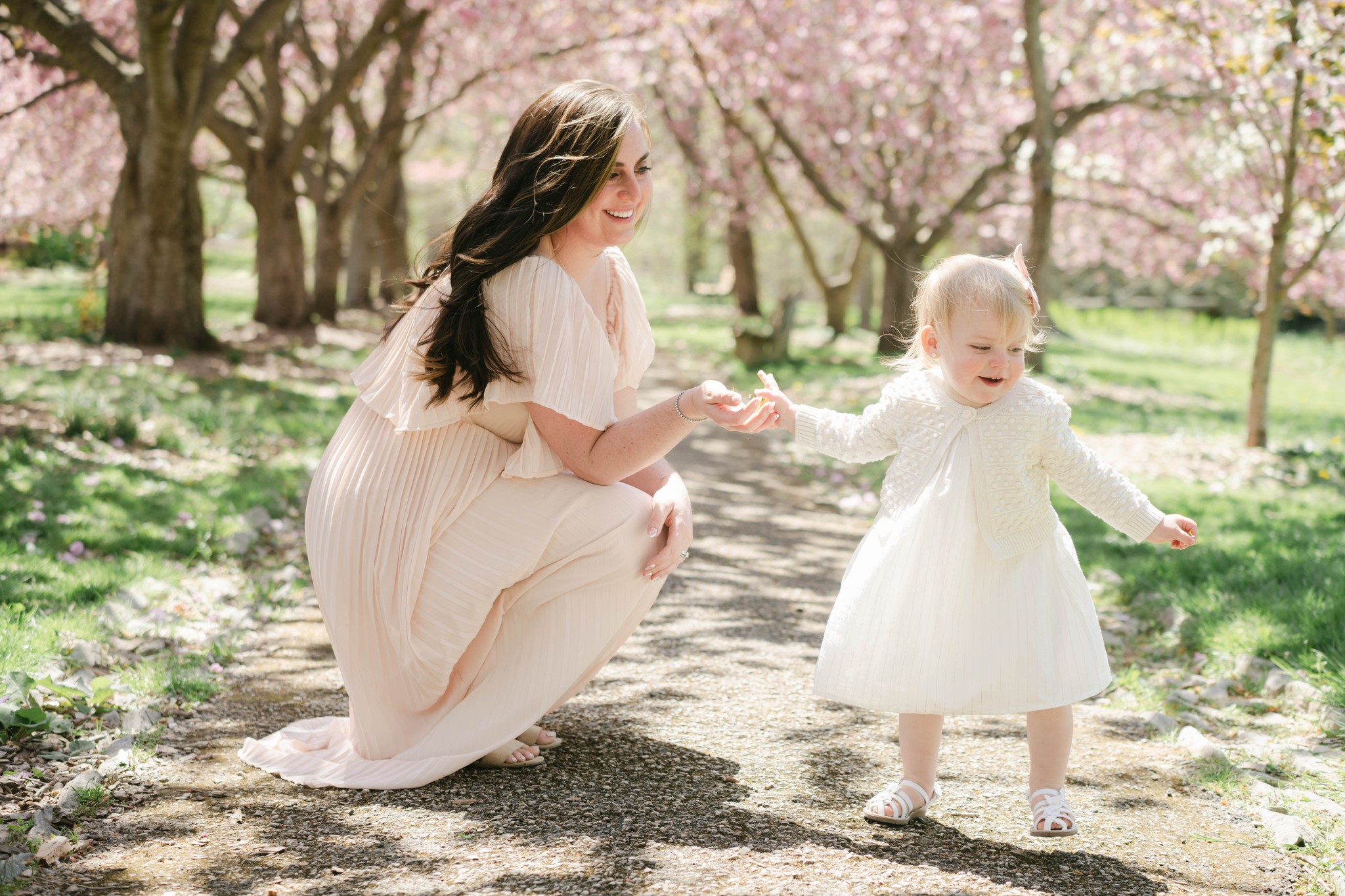 A mother plays with her toddler daughter in a white dress and sweater in a park path lined with trees blooming pink flowers luxey little ones